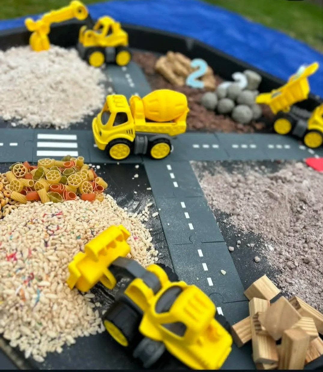 Toy construction vehicles and building materials arranged on a play road, including yellow toy dump trucks, piles of rice, pasta, and other grains, along with small wooden blocks and a model road with painted markings.