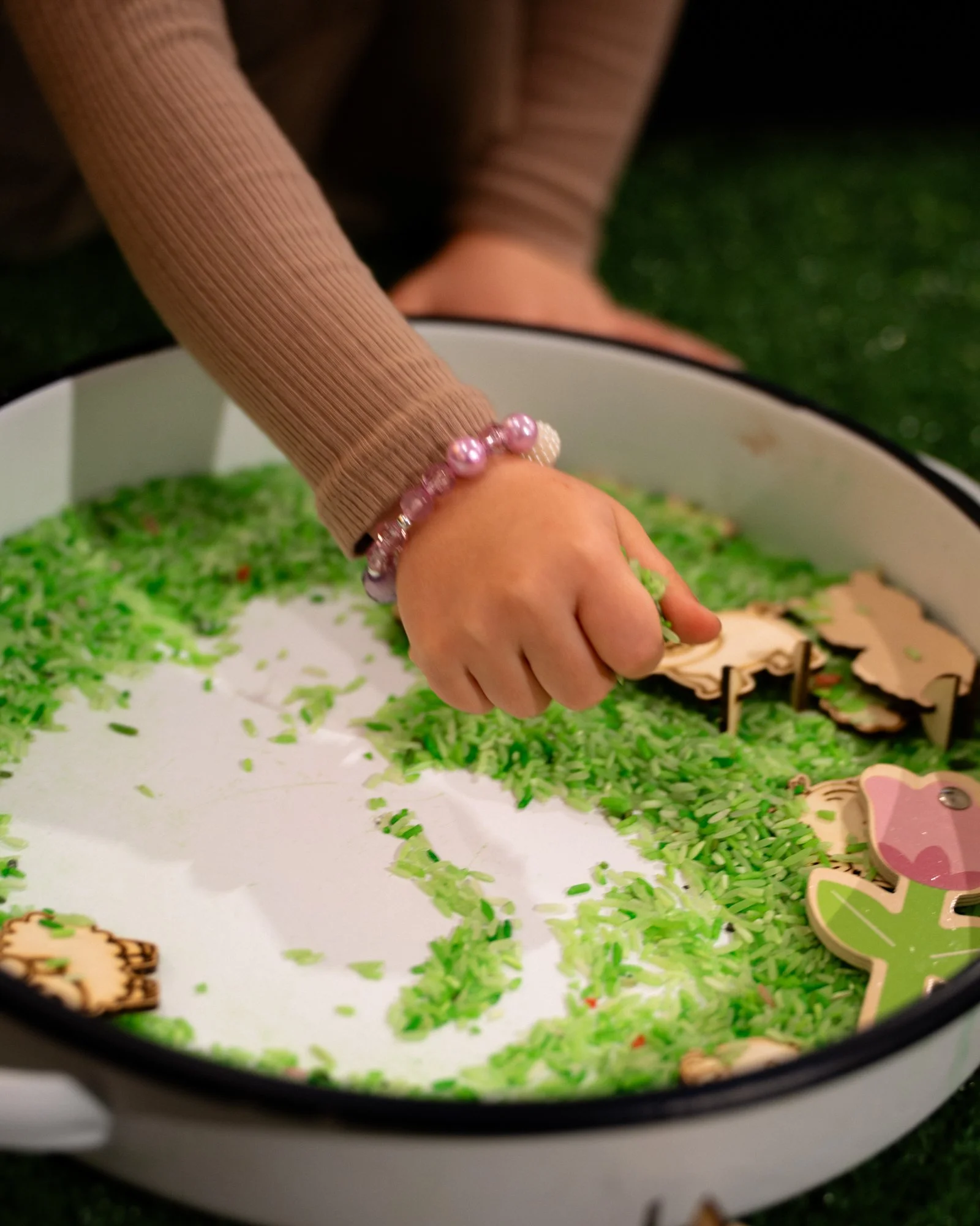 A child's hand reaching into a container of Christmas tree ground cover confetti, with some holiday cookies nearby. The child is wearing a beaded bracelet.