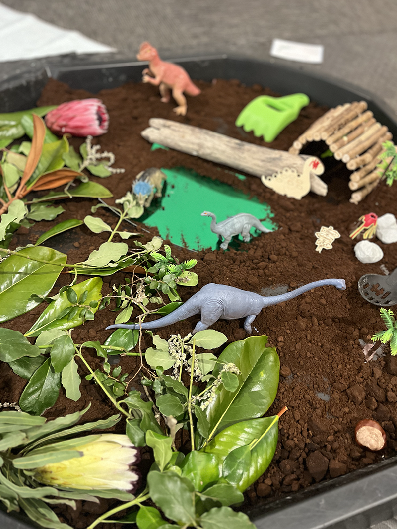 Toy dinosaurs and miniatures arranged in a miniature prehistoric scene on a dirt-filled surface with greenery and small accessories.