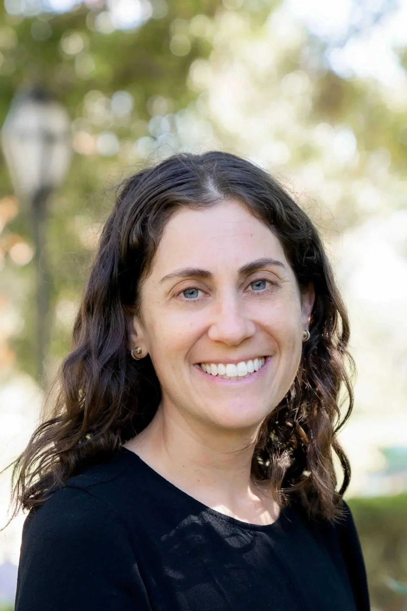 A smiling woman with shoulder-length curly brown hair, blue eyes, wearing a black top, standing outdoors with blurred trees and sunlight in the background.