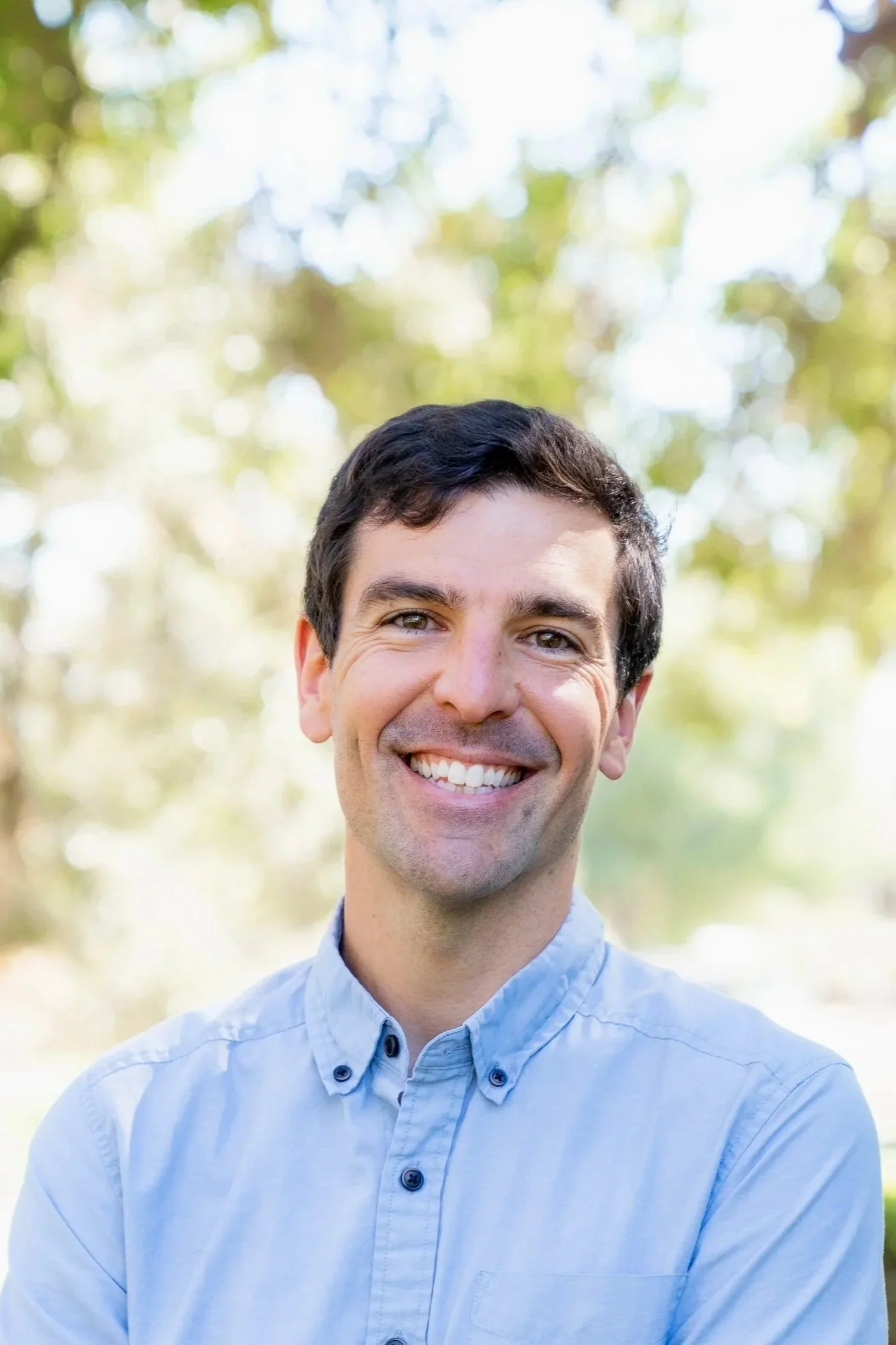 A smiling man with dark hair wearing a light blue button-up shirt outdoors, with a blurred background of trees and sunlight.
