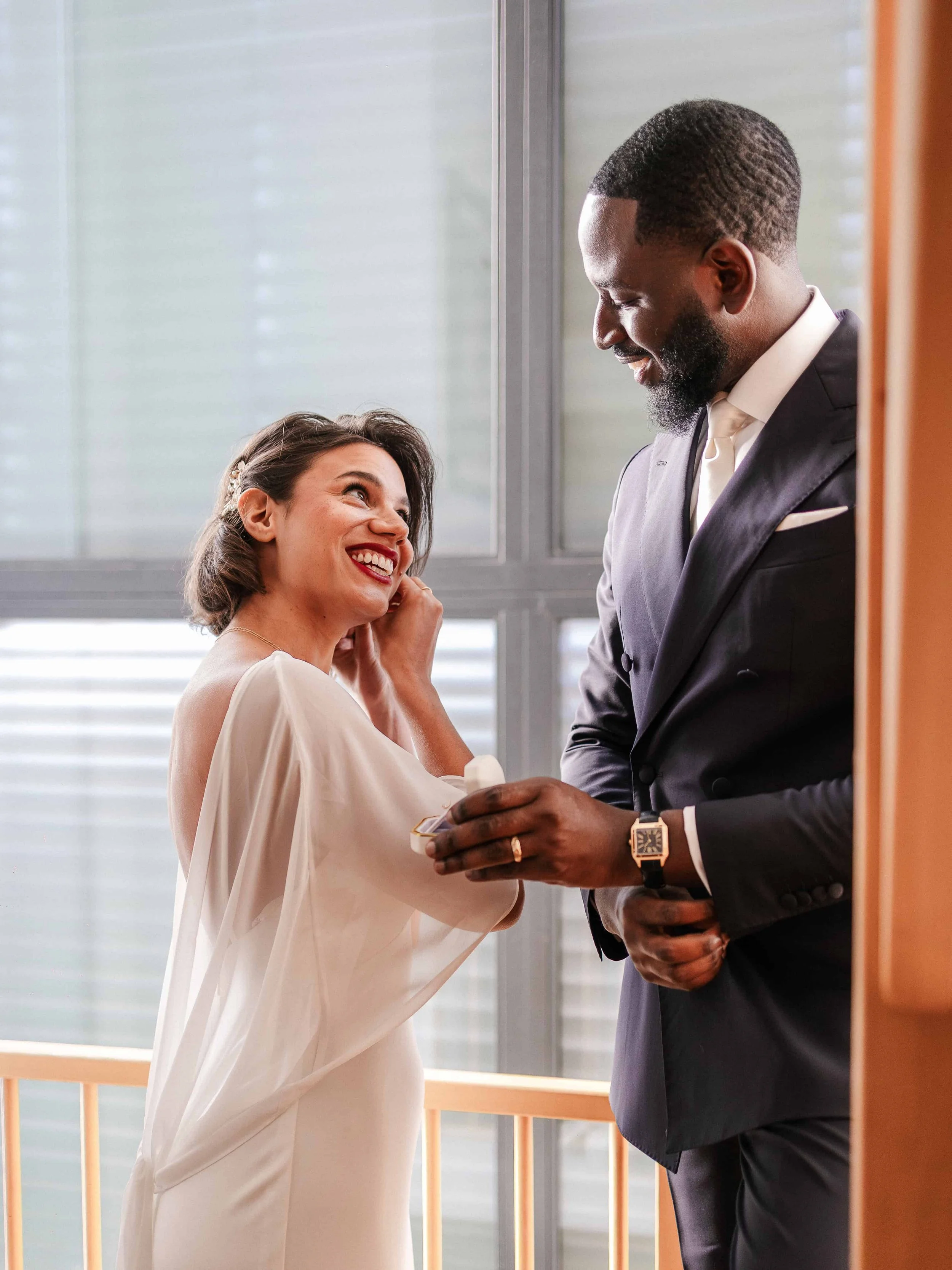 Een glimlachende vrouw in een witte jurk ontvangt een ring van een man in een zwart pak, beide glimlachend en makend oogcontact.