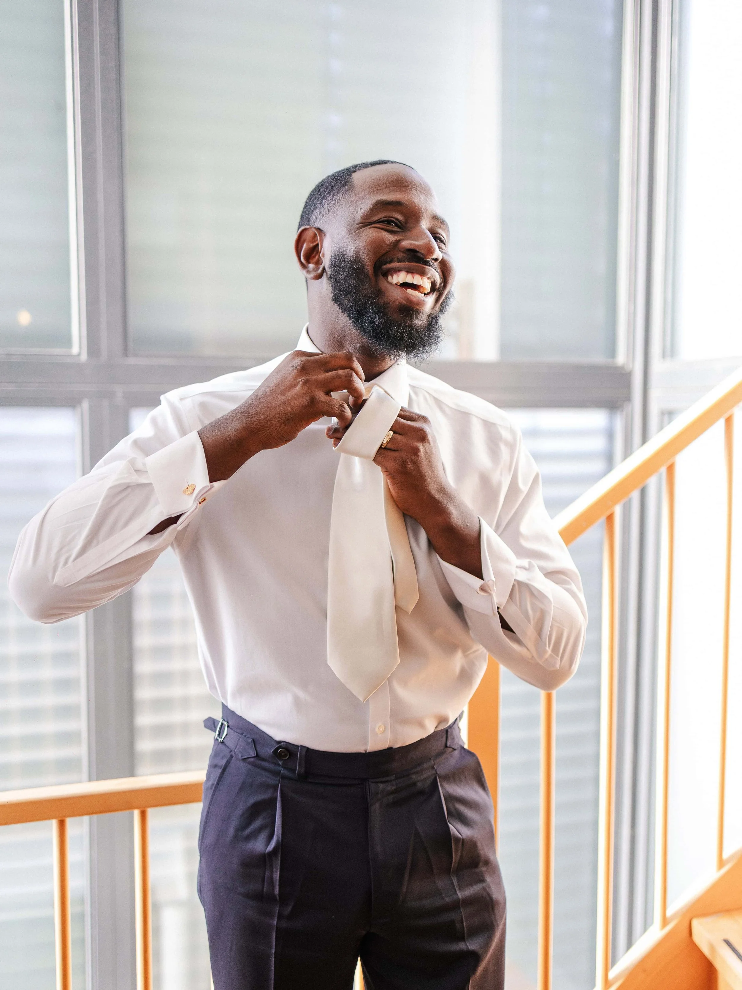 Man in wit overhemd en donker pak, die een stropdas knoopt, glimlachend in een lichte kamer met grote ramen.