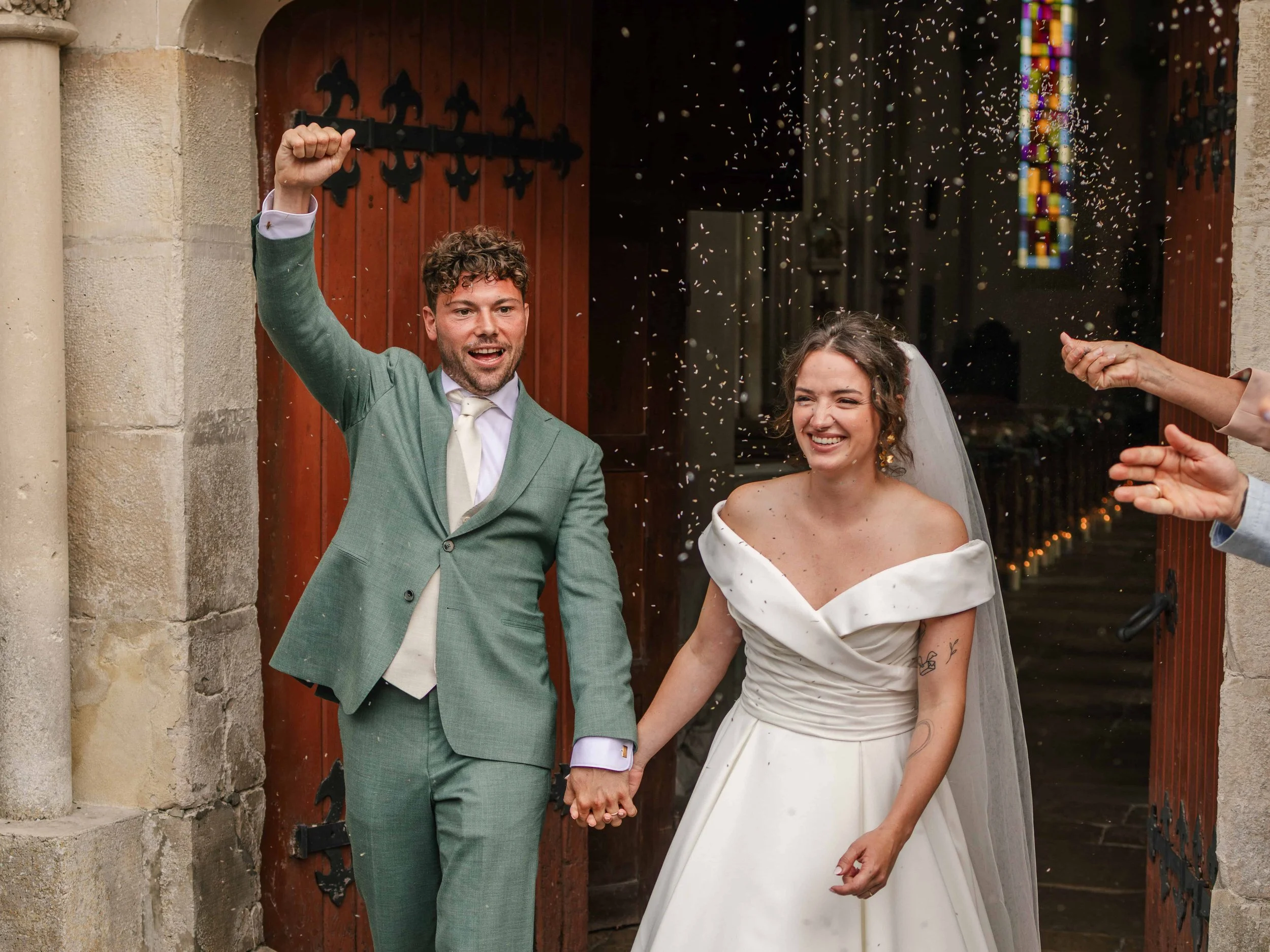 Bruidegom in groen pak en bruid in witte jurk die hand in hand buiten een kerk uitlopen, terwijl confetti wordt uitgestrooid.