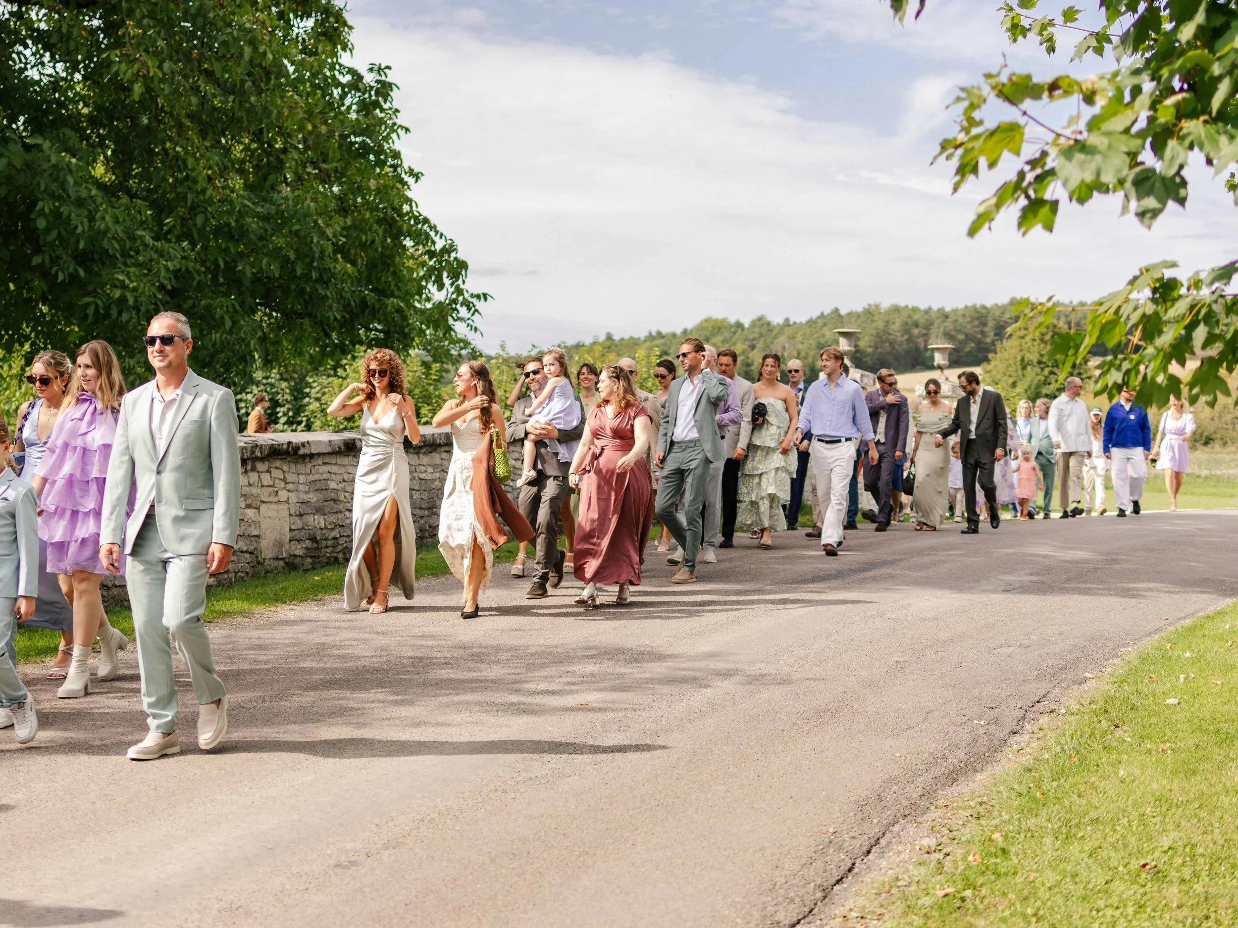 Mensen die in een rij lopen op een pad in een buitenomgeving, mogelijk tijdens een feest of bruiloft, met veel groen en een grijze stenen muur op de achtergrond.