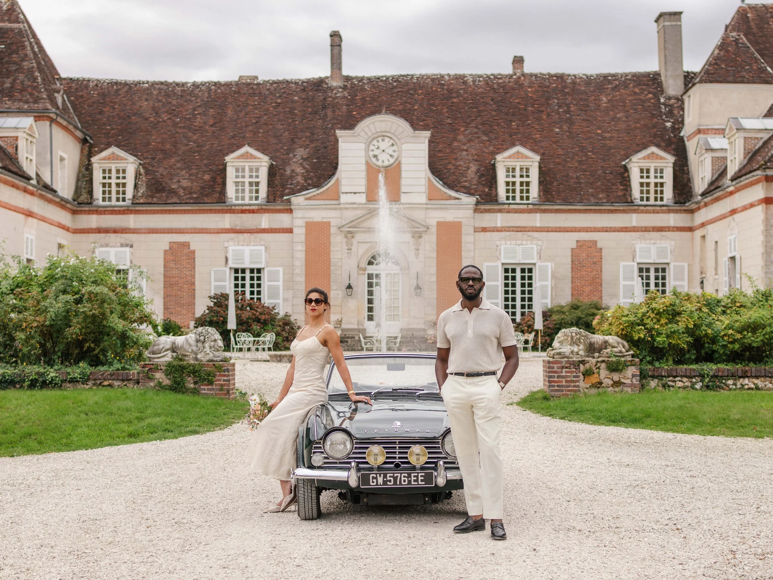 Een man en een vrouw poseren voor een historaal landhuis met een klassieke auto op de voorgrond. De vrouw draagt een crèmekleurige jurk en de man een lichtkleurige broek en polo. Het landschap is mooi en goed onderhouden met groene struiken en beelde