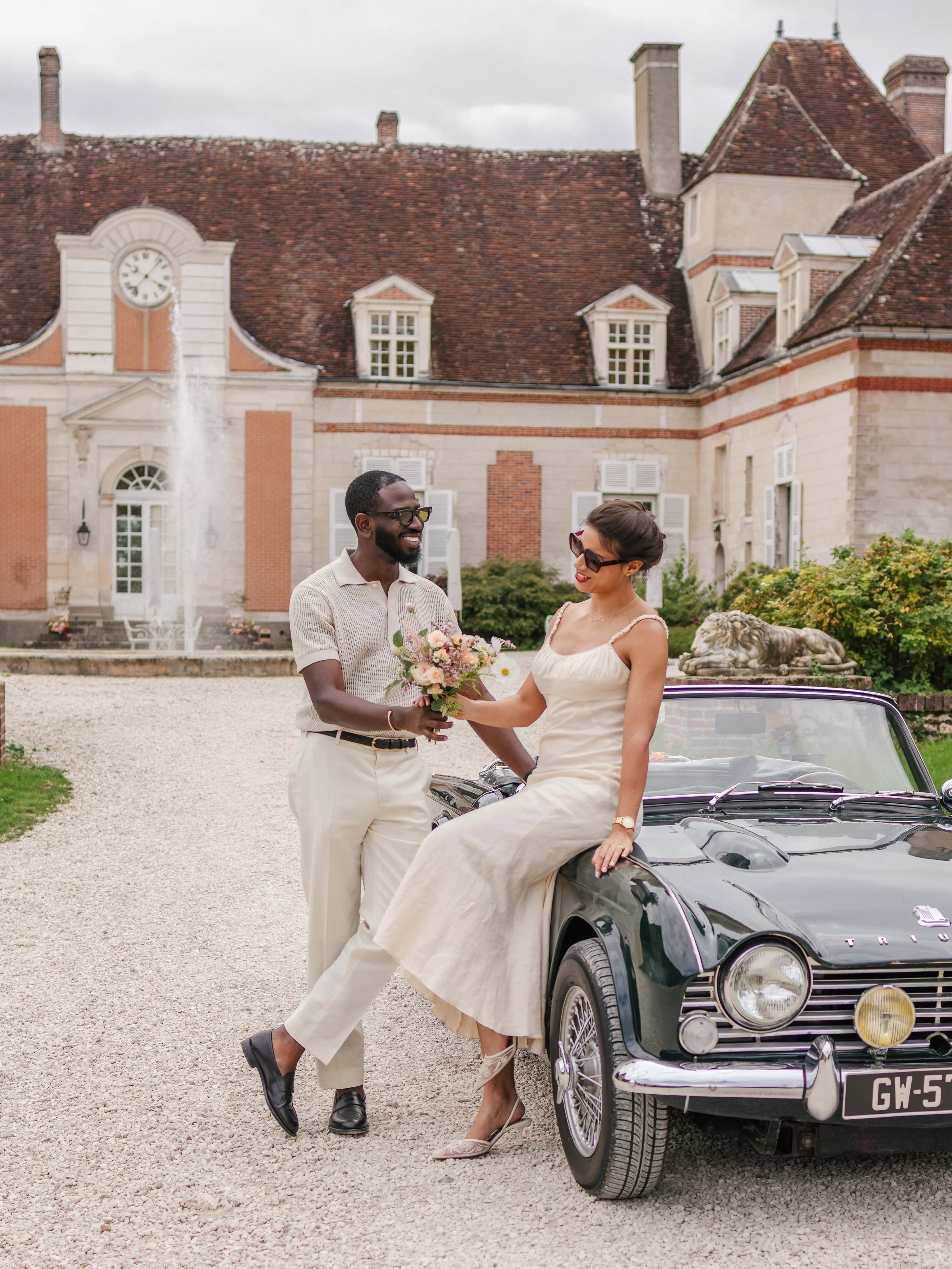 Een zwarteman en een blanke vrouw op een vintage auto voor een landhuis, zij ruilt een bloemensamenstelling uit.