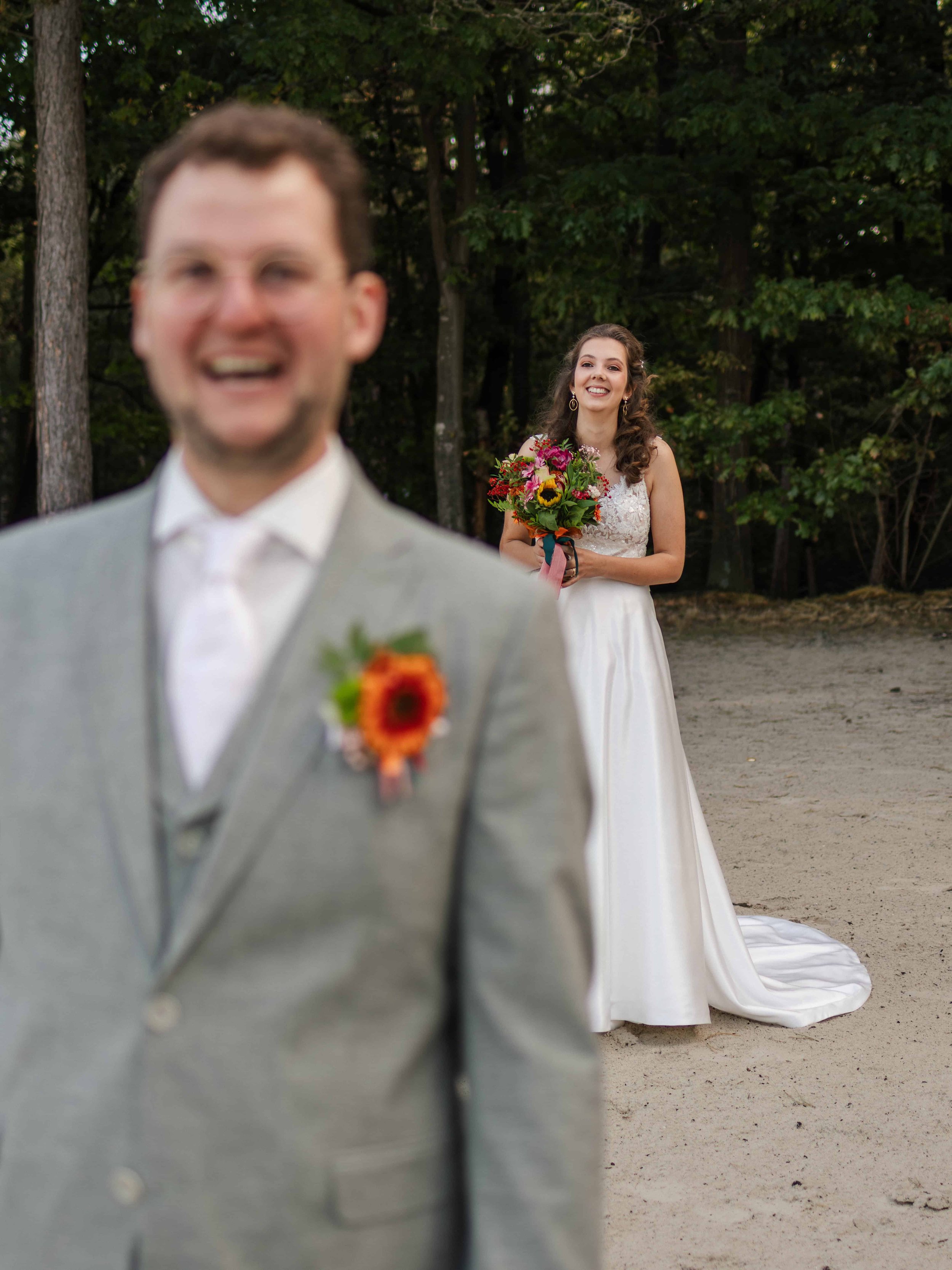 Bruidegom in de voorgrond lachend, vage afbeelding, en bruid op de achtergrond met een boeket bloemen, buiten op een zandterrein, met een achtergrond van bomen, tijdens een bruiloft.