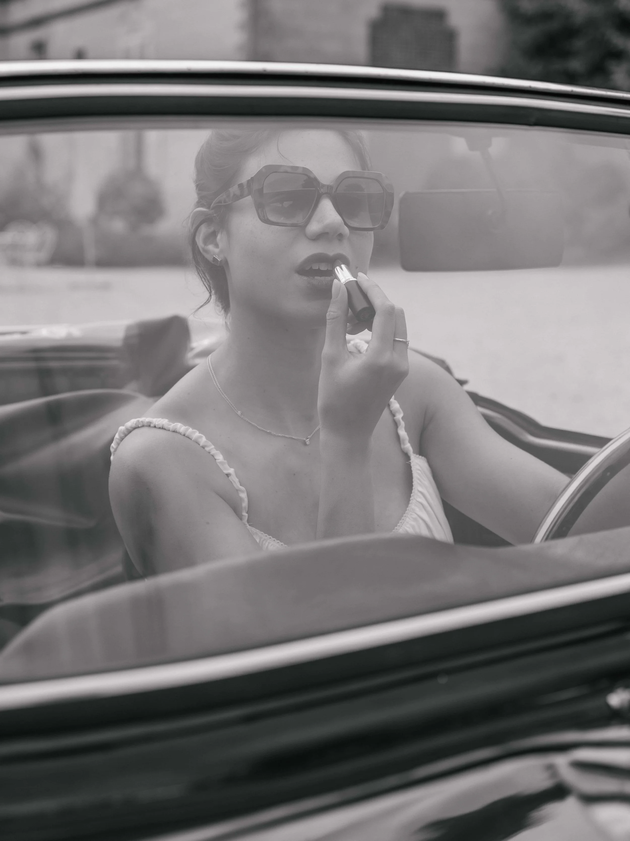 Een vrouw met grote zonnebril die lipstick aanbrengt in een auto, gezien door de voorruit.