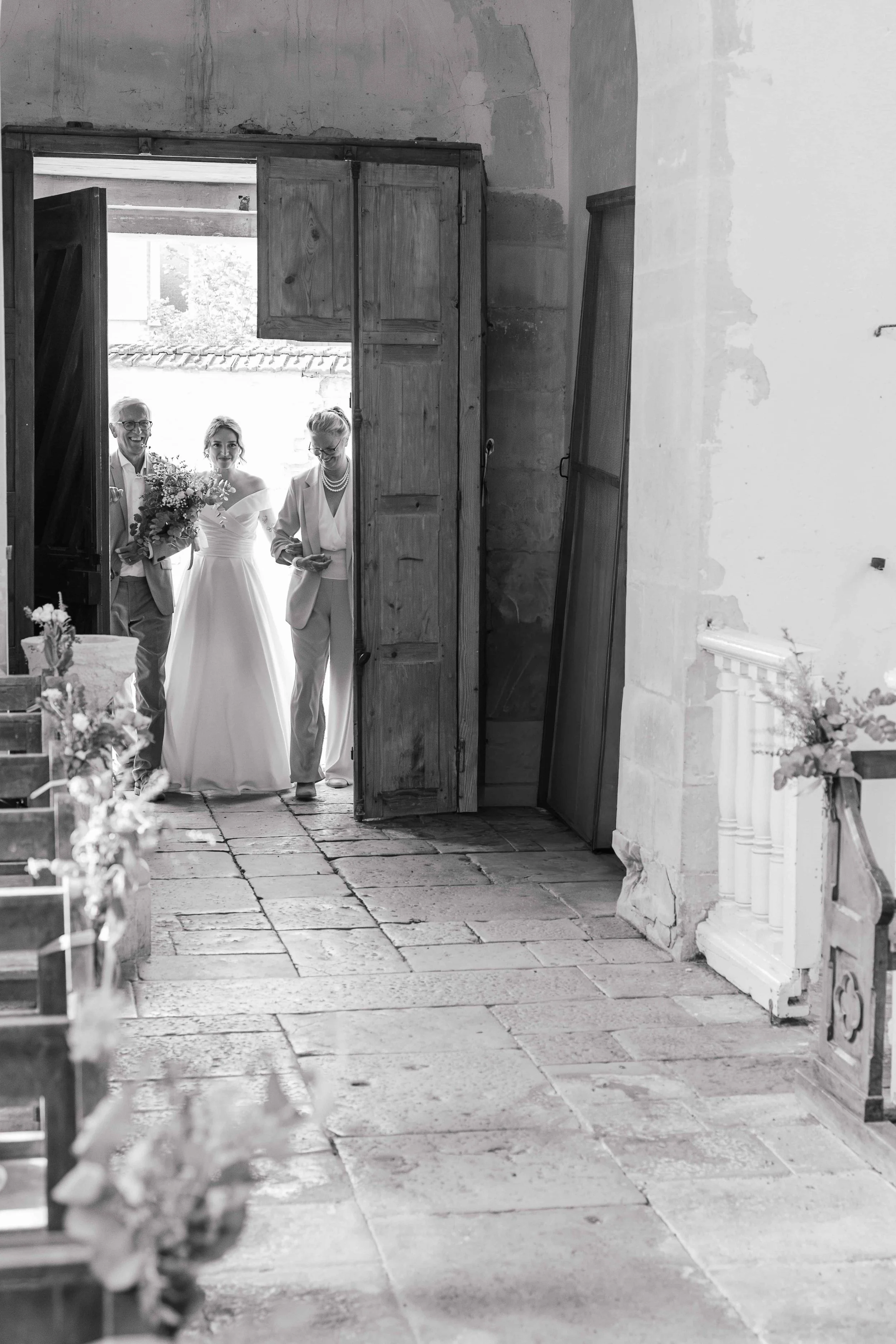 De foto toont een bruiloft in een oude stenen kerk met een houten deur. Een bruid met een witte jurk en een bouquet komt naar buiten, vergezeld door twee mensen, mogelijk familie. Ze lijken blij en opgewonden.