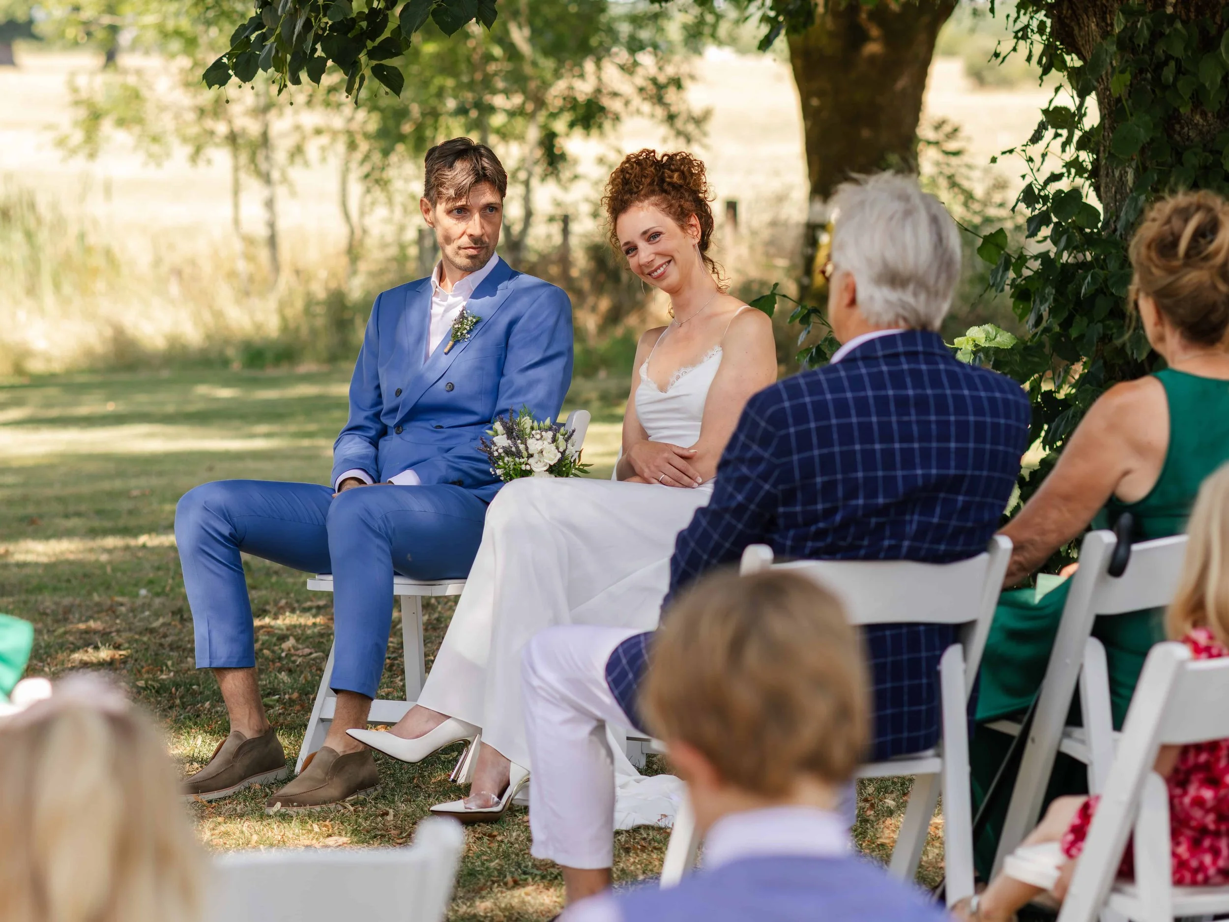 Een bruid en een bruidegom zitten tijdens een buitenhuwelijk, omringd door gasten onder een boom op een grasveld, met een landelijke achtergrond.