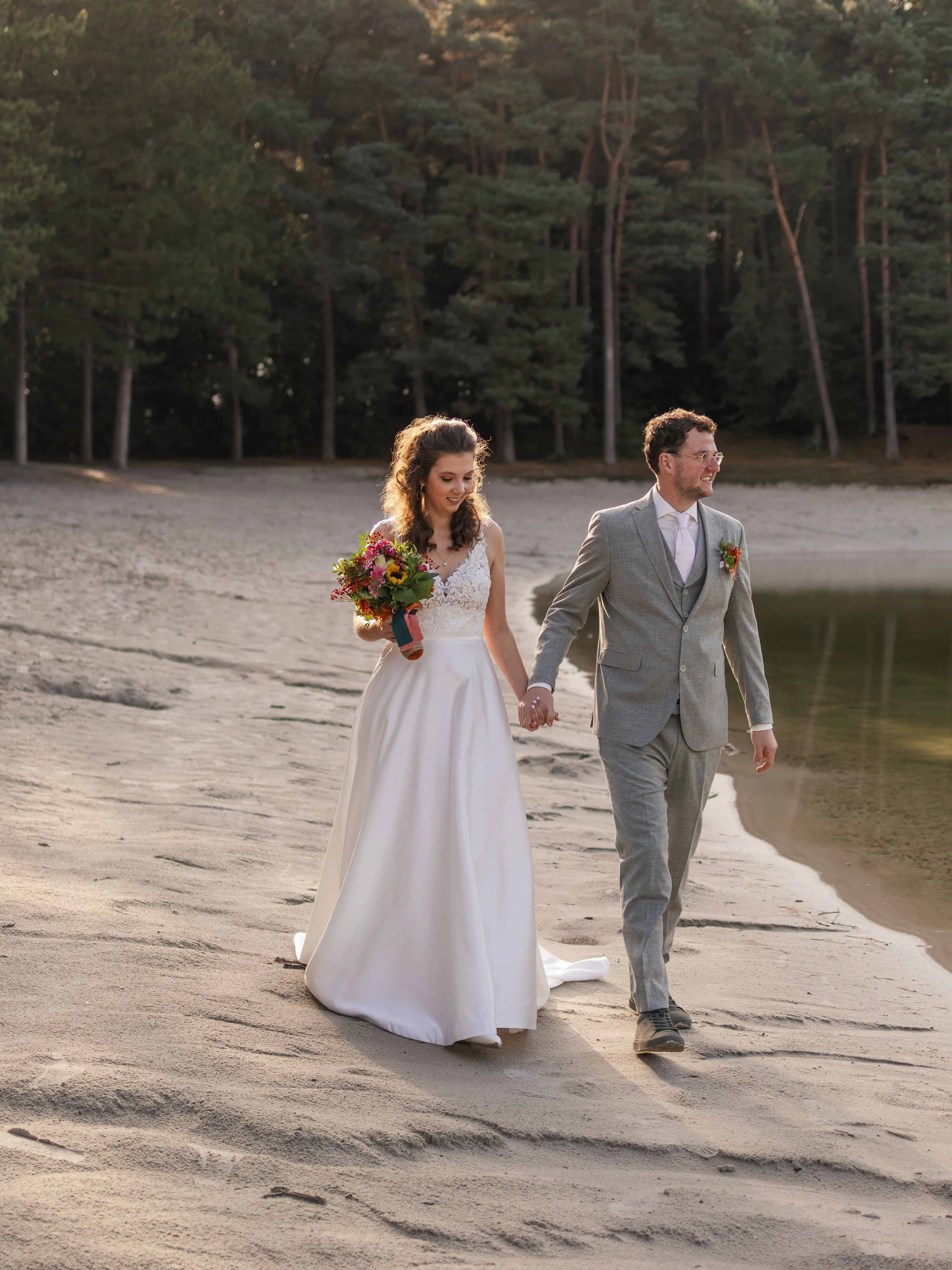 Een bruid en bruidegom lopen hand in hand langs de oever van een meertje of rivier, omringd door bomen, tijdens een huwelijk. De bruid draagt een witte jurk en houdt een bloemboeket, terwijl de bruidegom een grijze pak draagt.