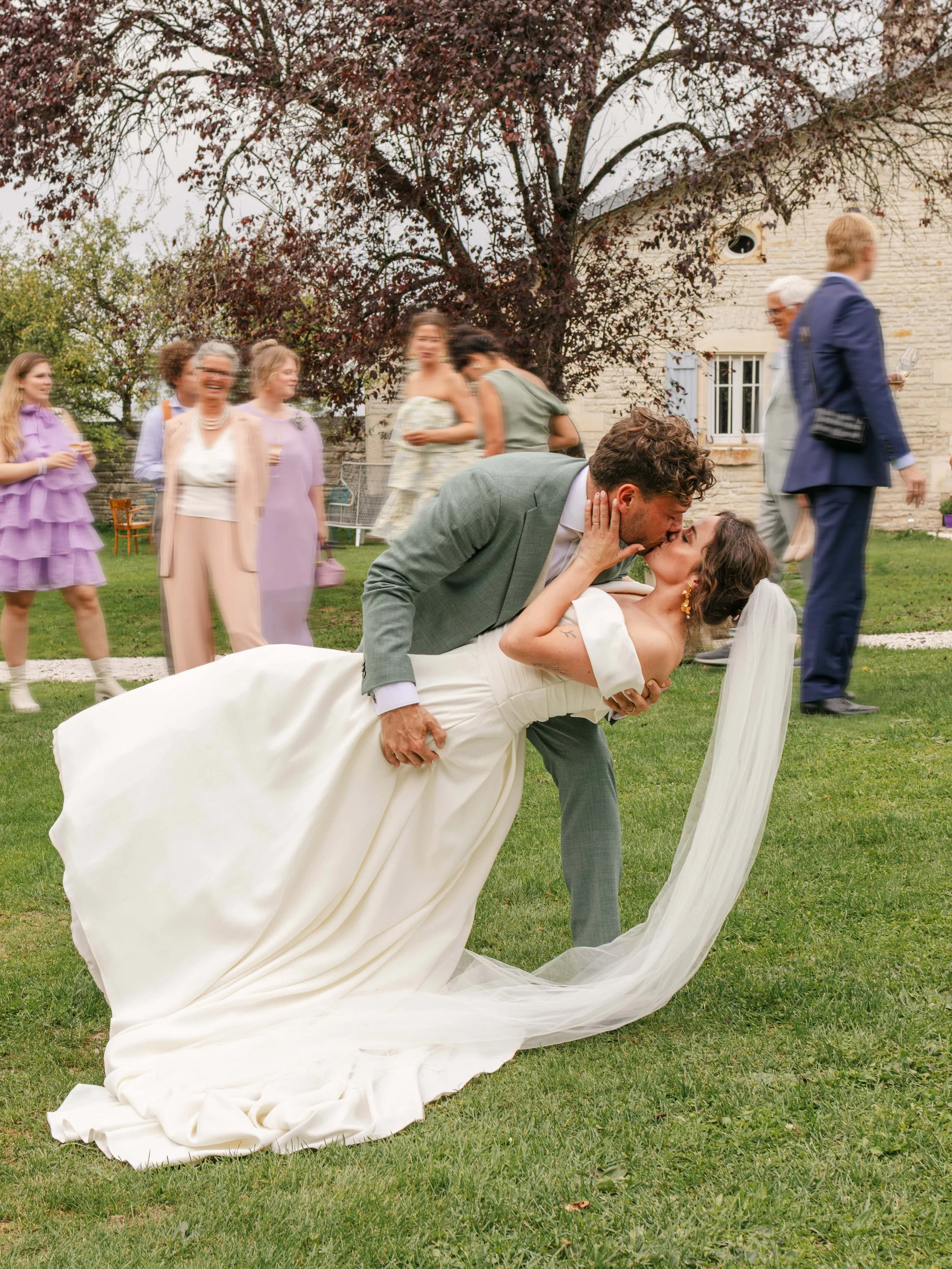 Koppel die elkaar zoent tijdens een bruiloftsfeest op een grasveld met gasten op de achtergrond.
