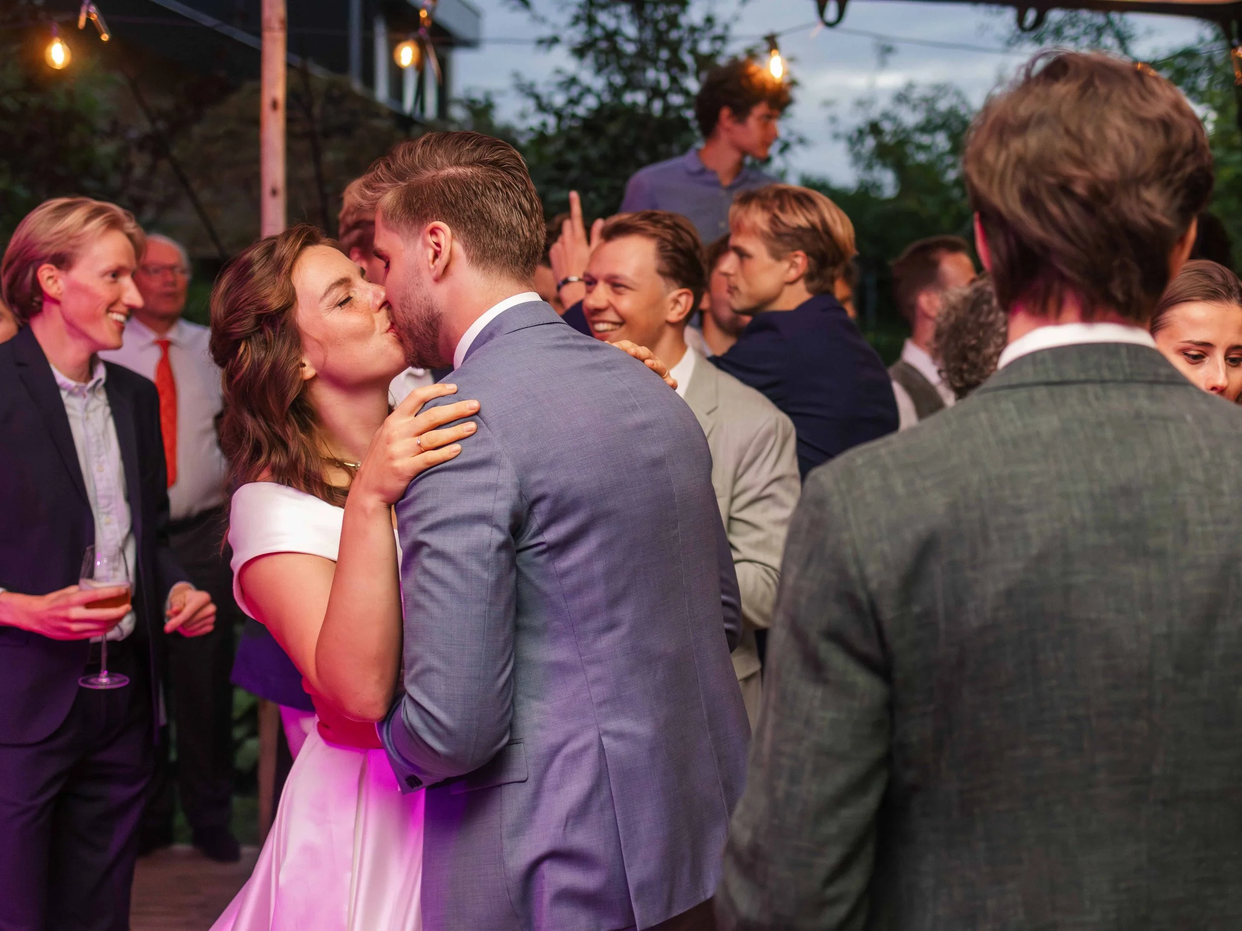 Een groep mensen op een feest of evenement, met een paar die kussen, onder feestverlichting en groene omgeving.