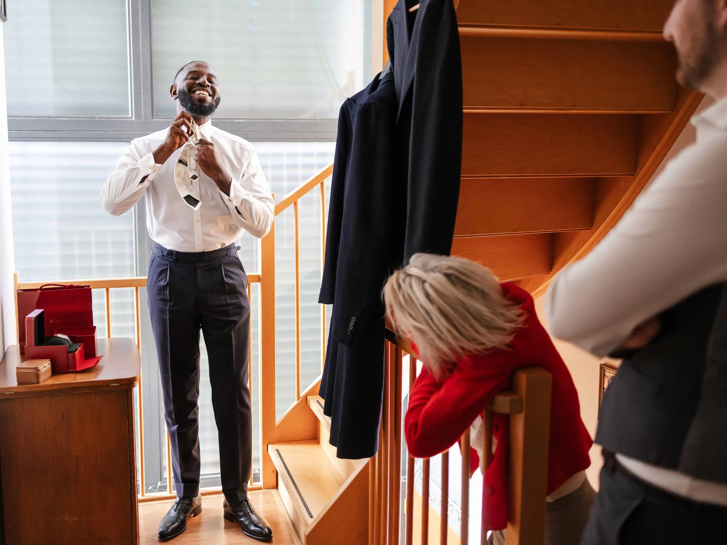 Man die een stropdas vastmaakt, anderen kijken toe, in een woning met trap en een grote raampartij.