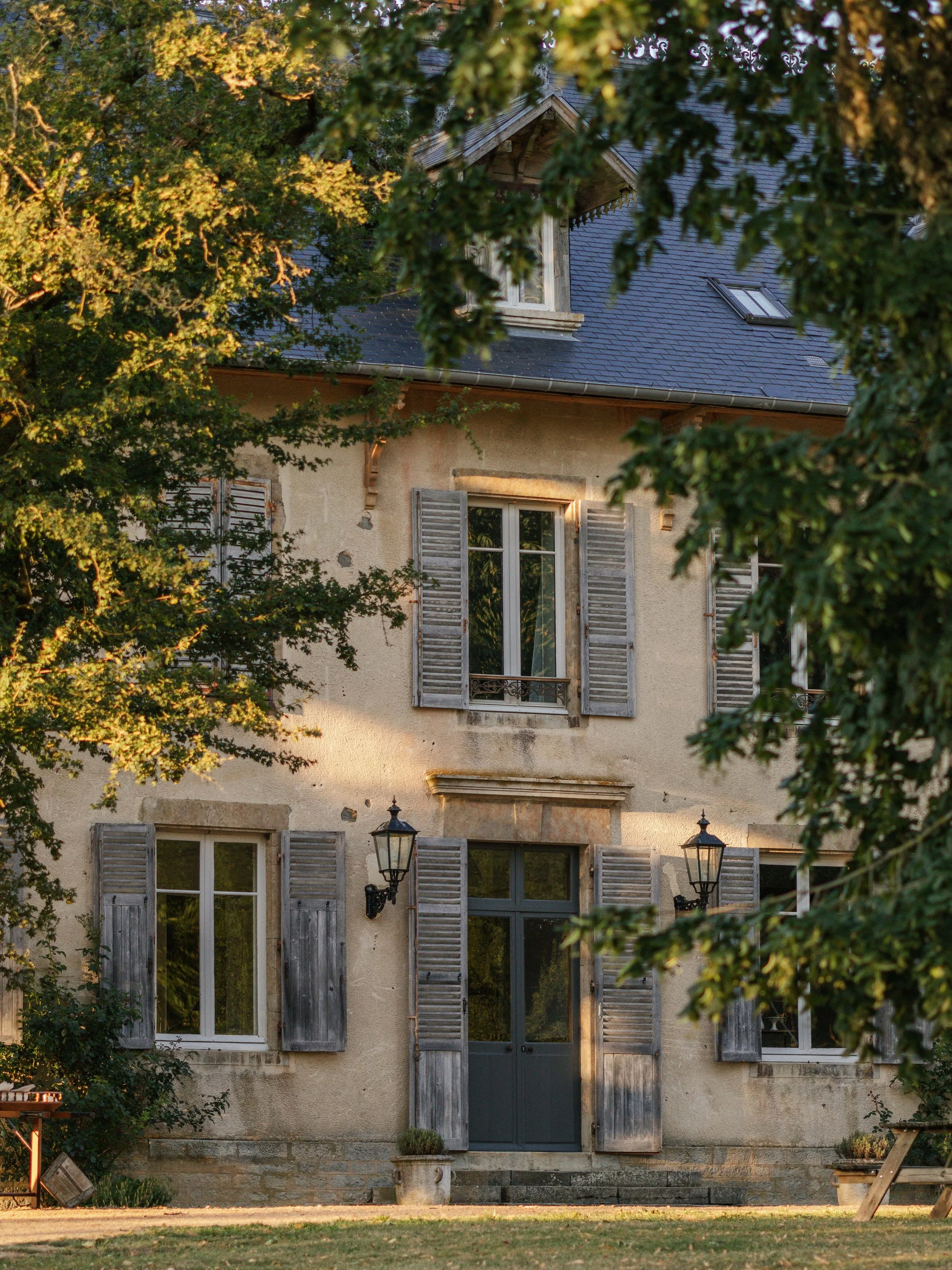 Een oude landelijke huisgevel met ramen en luiken, omgeven door bomen en tuin, in de avondzon.