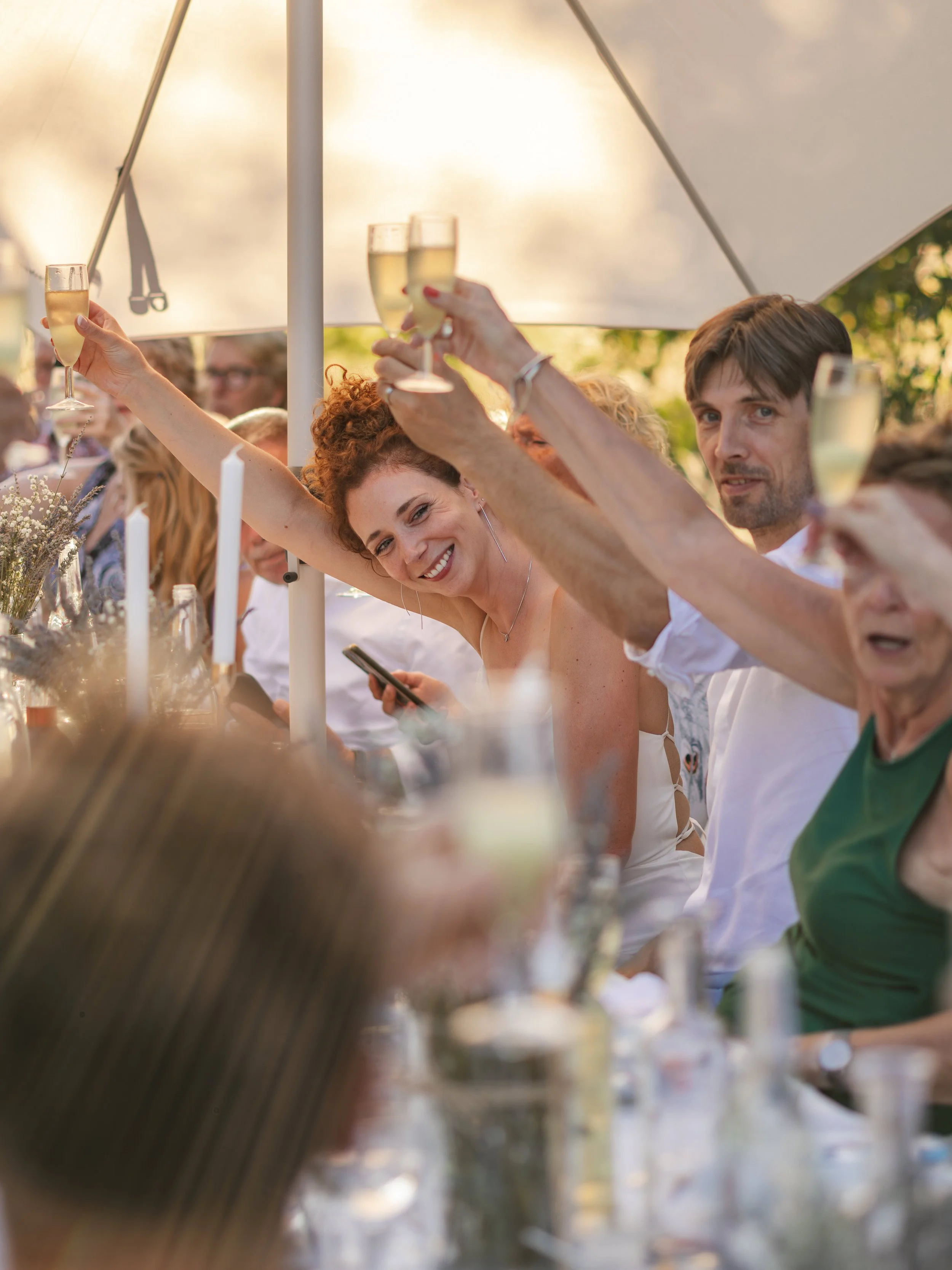 Mensen vieren een feestje onder een grote parasol, ze heffen glazen champagne en lachen, met een tafel vol drankjes en decoraties.