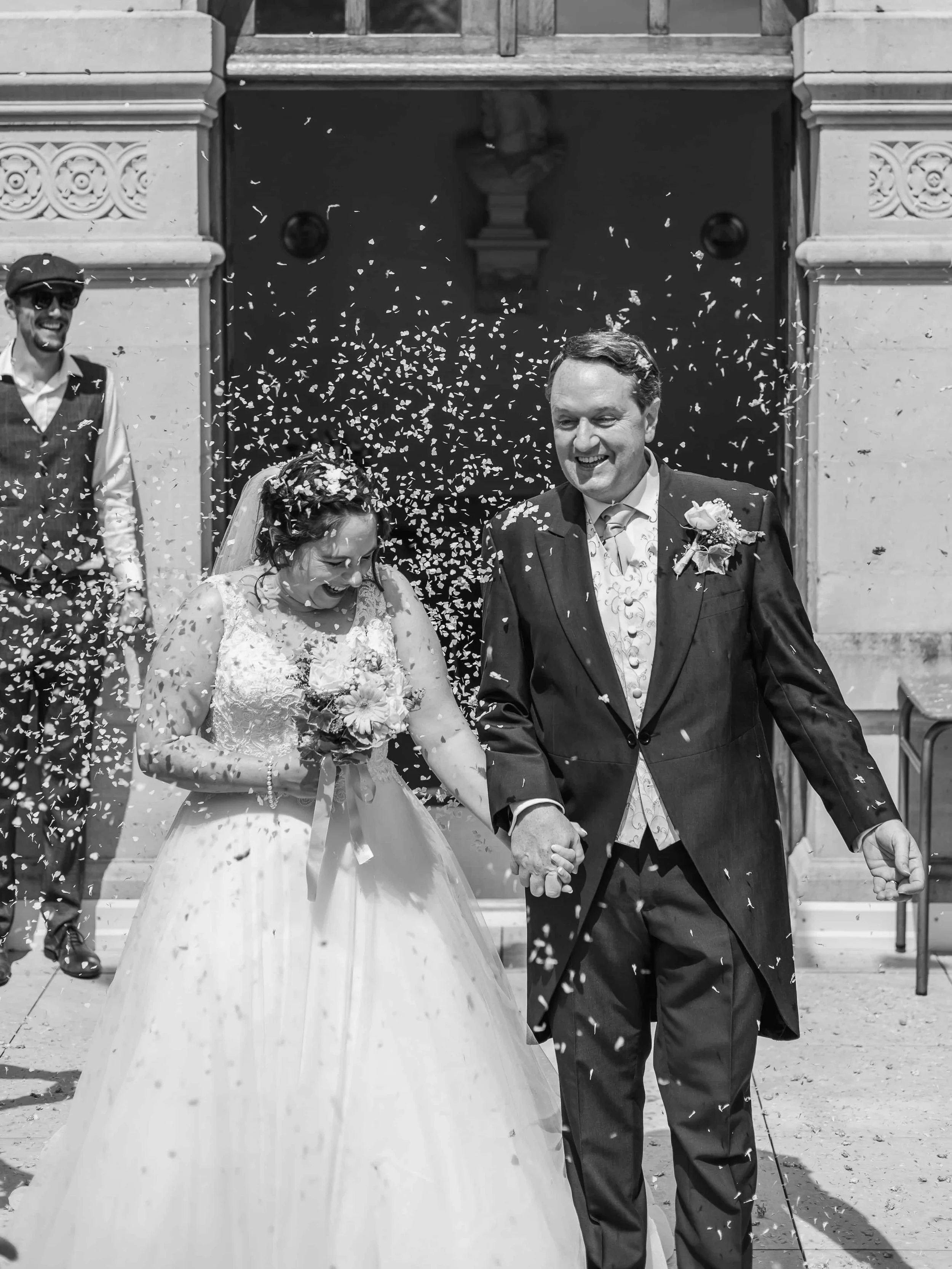 Bruidegom en bruid lopen hand in hand onder confetti na hun huwelijksceremonie, in zwart-wit.