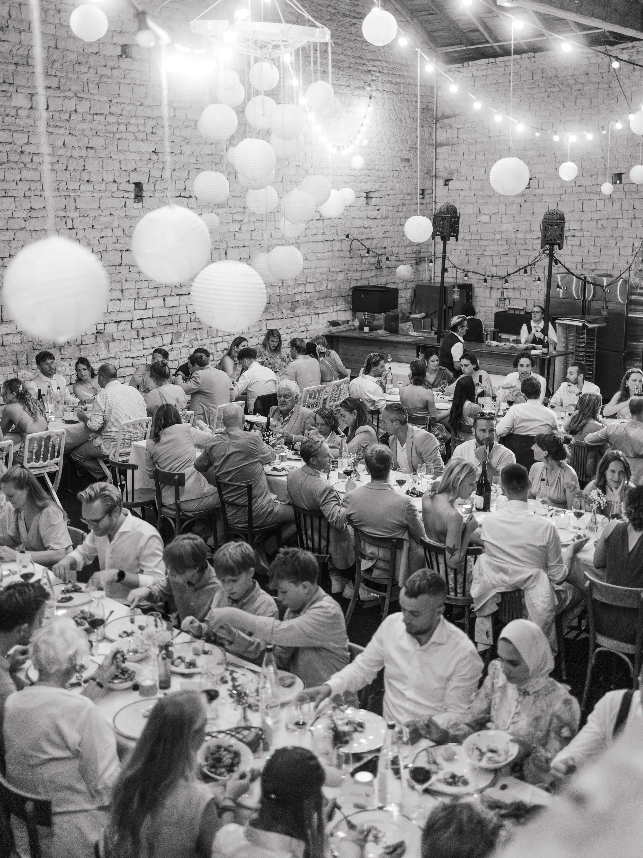 een grote feestzaal met veel mensen die aan lange diners genieten, decoratie met witte papieren lantaarns en stringverlichting, en een DJ booth op de achtergrond.