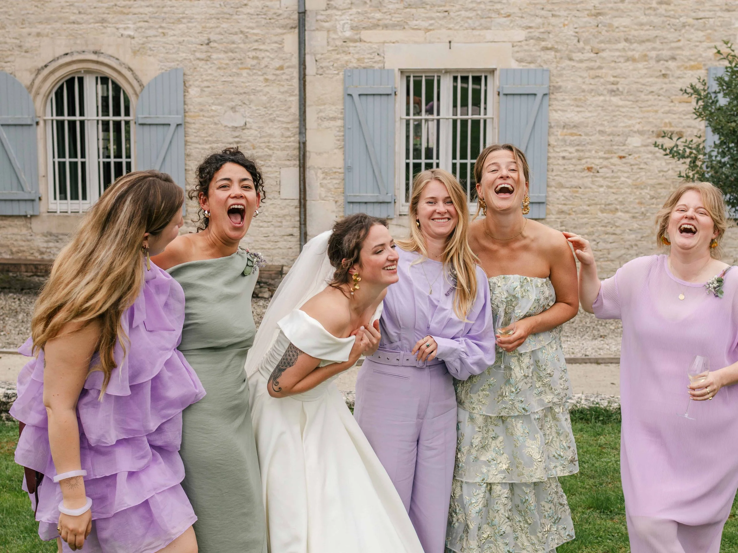 Vijf vrouwen en een bruid lachen en genieten op een buitenfeest, met een stenen huis op de achtergrond.