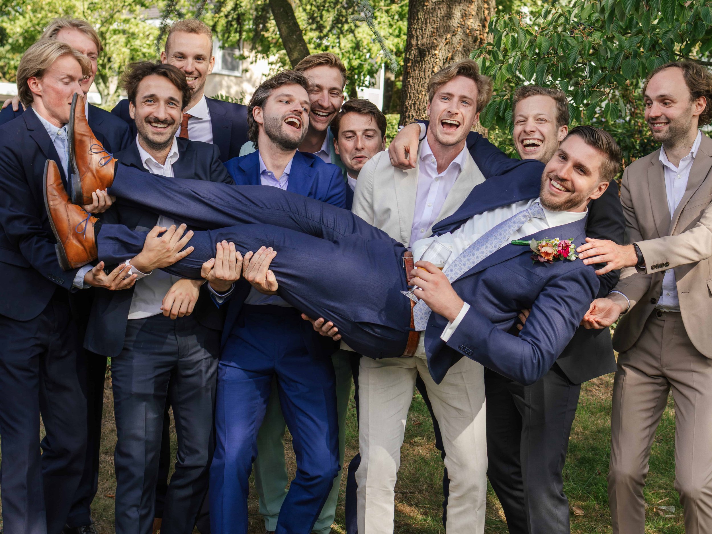 Group van vrolijke mannen in formele kleding, die een man in een donkerblauw pak en gestreept lichtblauw shirt optillen tijdens een feestje of bruiloft in een tuin met bomen.