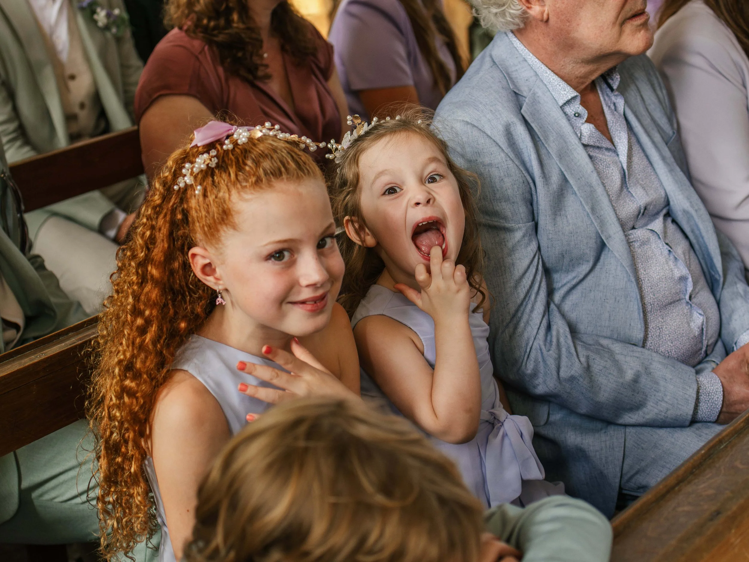 Twee jonge meisjes en een oudere man zitten op een bank in een kerk, de meisjes dragen bloemenkransen en jurkjes, een van de meisjes lacht en wijst naar de camera terwijl de andere zich vrolijk uitdrukt.