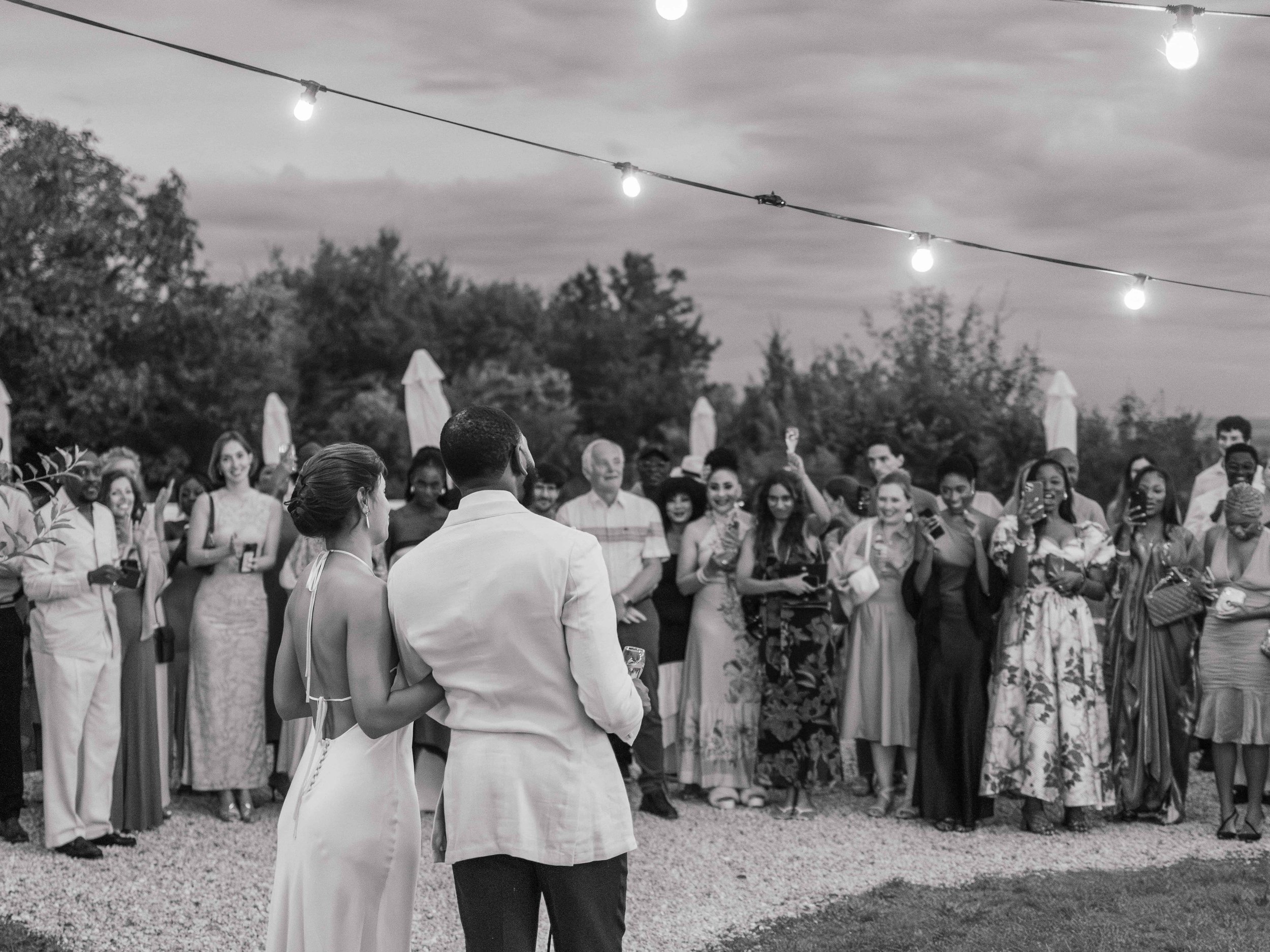 Een zwart-wit afbeelding van een bruiloft buitenshuis met een groot groep mensen die naar het bruidspaar kijkt. Er hangen lampjes boven de menigte en de achtergrond heeft bomen en een bewolkte hemel.