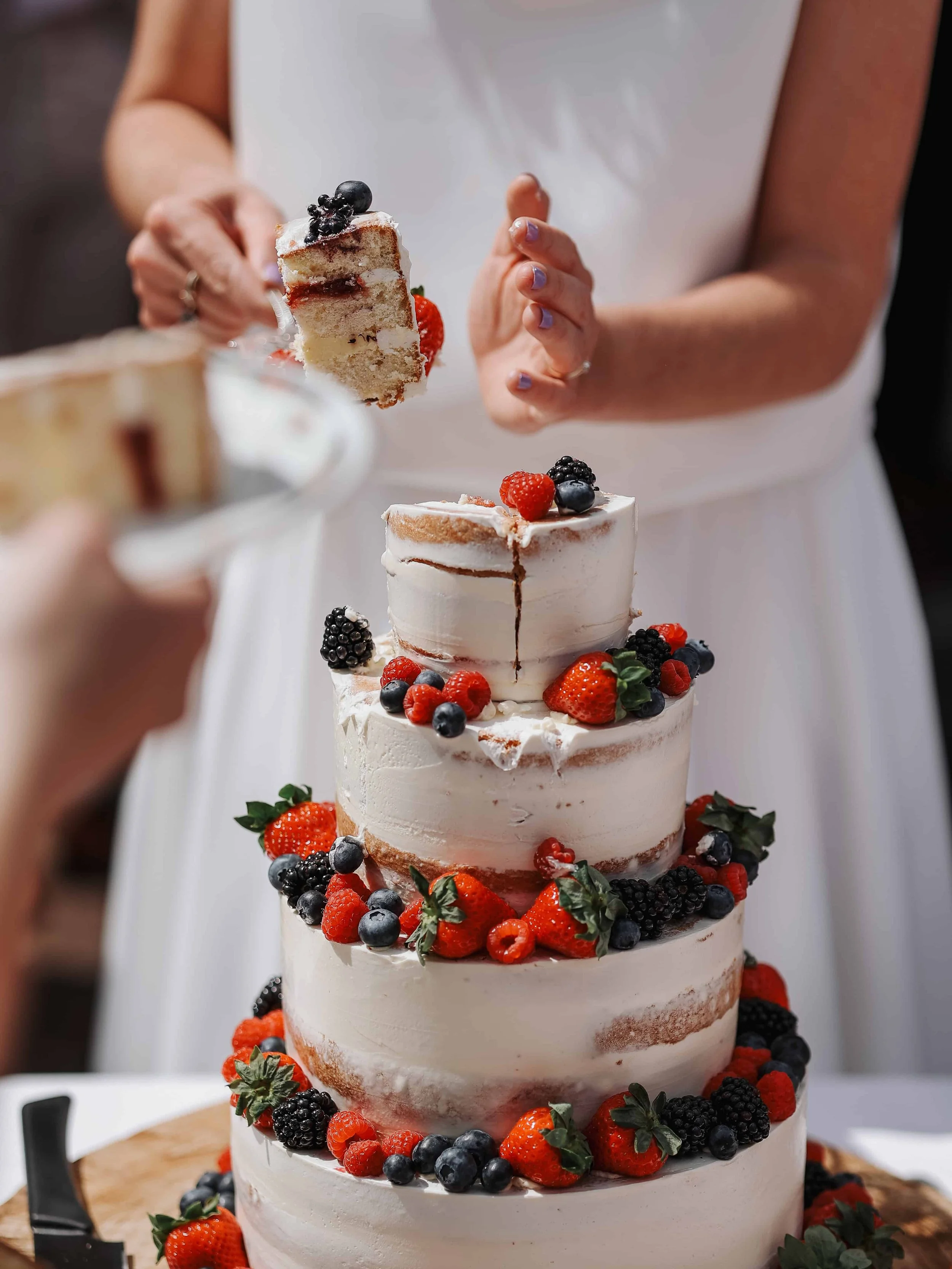 Een persoon snijdt een stuk taart van een vierlaagse bruidstaart die versierd is met gemengde bessen zoals aardbeien, bramen en blauwe bessen.
