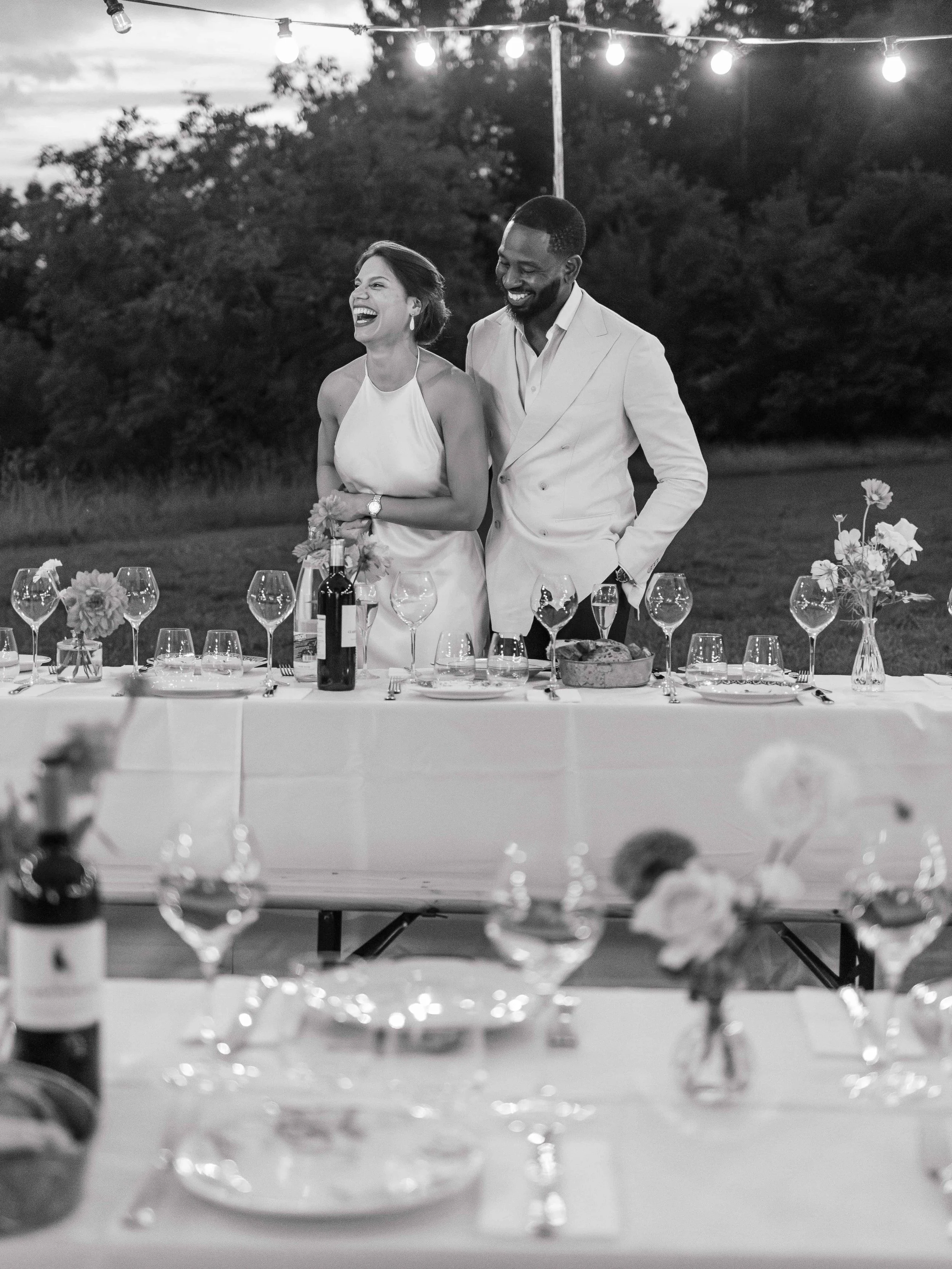 Een bruid en bruidegom staan samen achter een feestelijke dinertafel buiten, omringd door vorken, wijnglazen en bloemen, tijdens een avondfeest onder sfeervolle lichtjes.