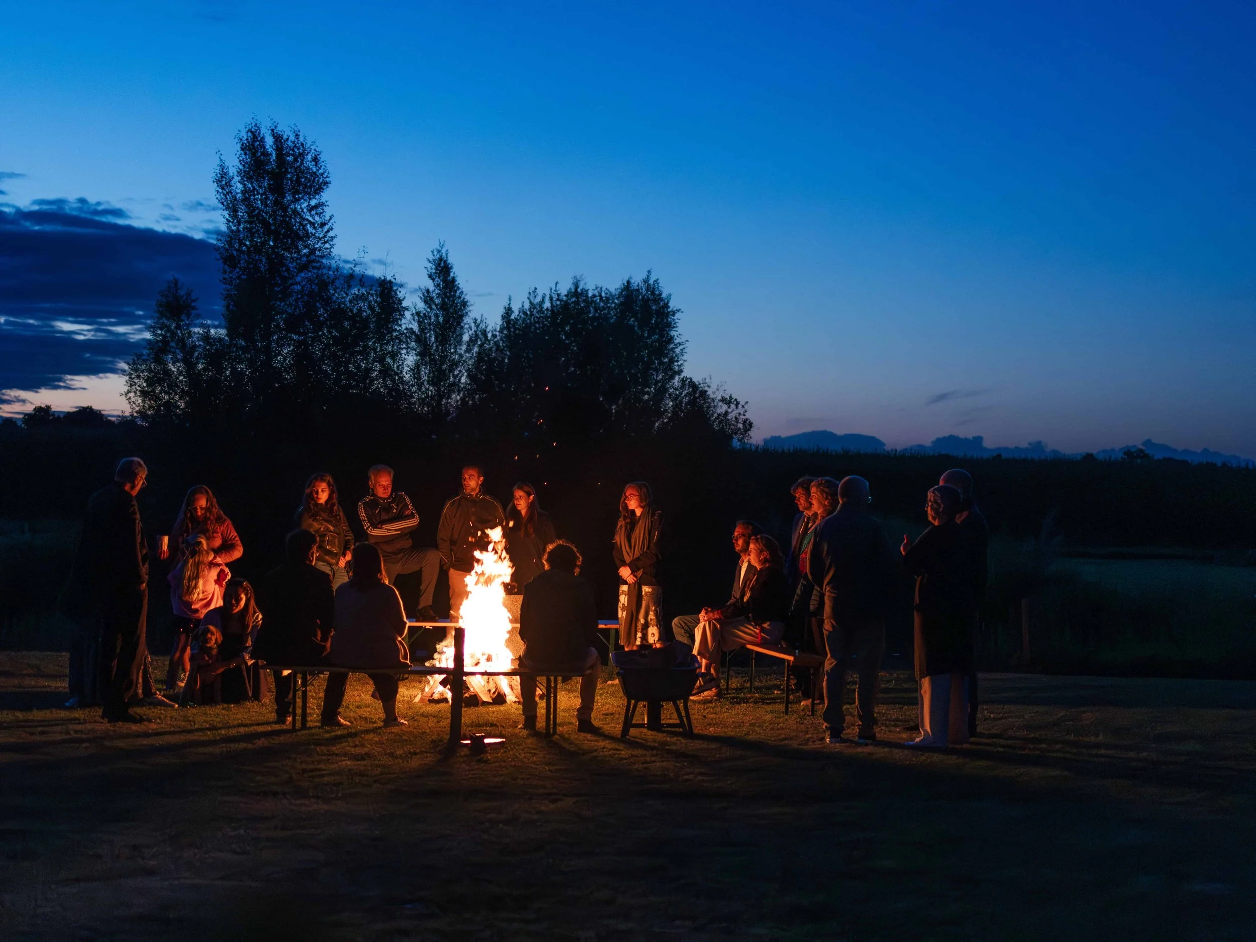 Groep mensen die bij een kampvuur zitten en eromheen staan tijdens de schemering in een open veld met bomen in de achtergrond.