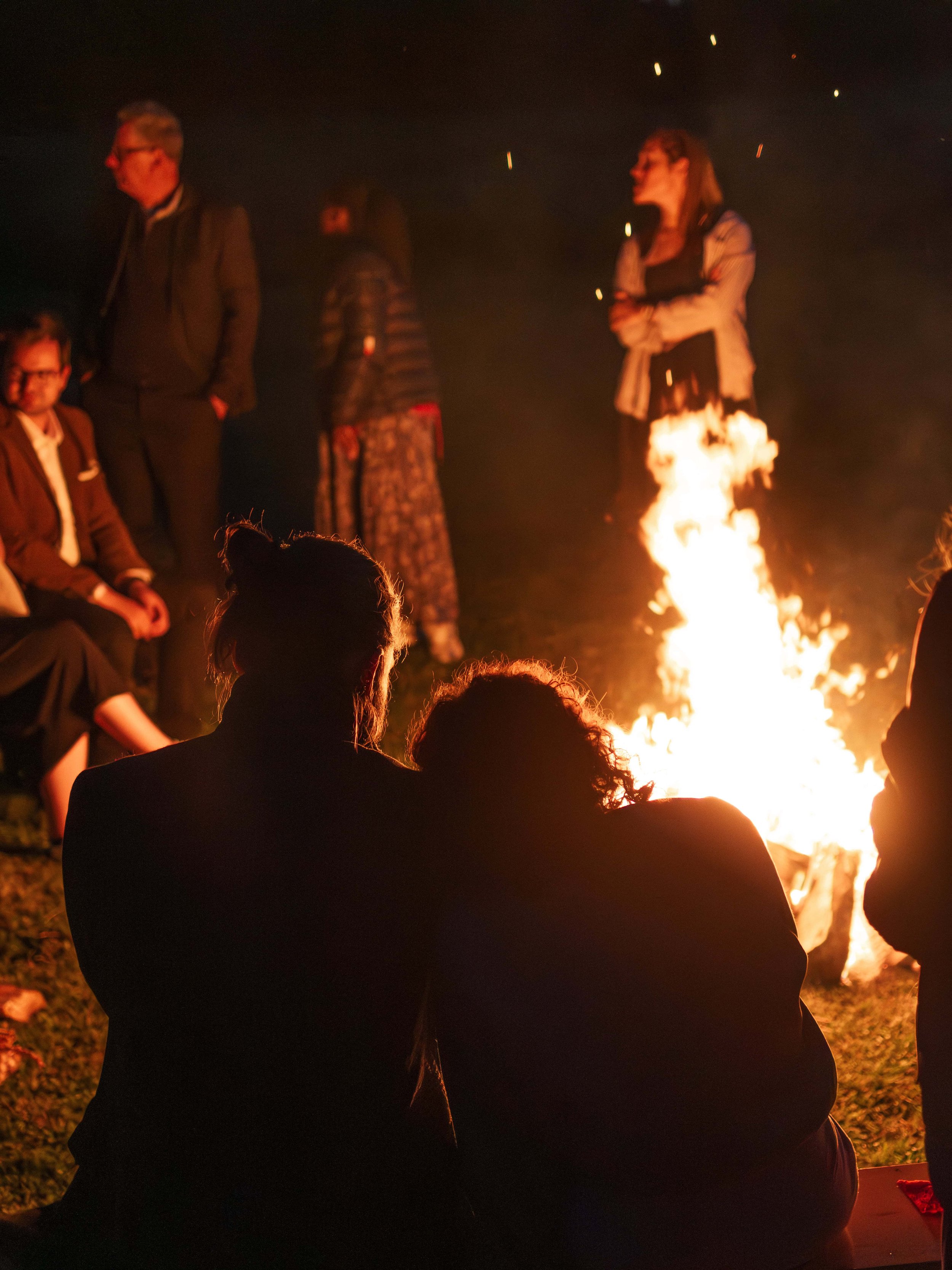 Mensen zitten rond een kampvuur 's nachts, enkele mensen staan en kijken toe