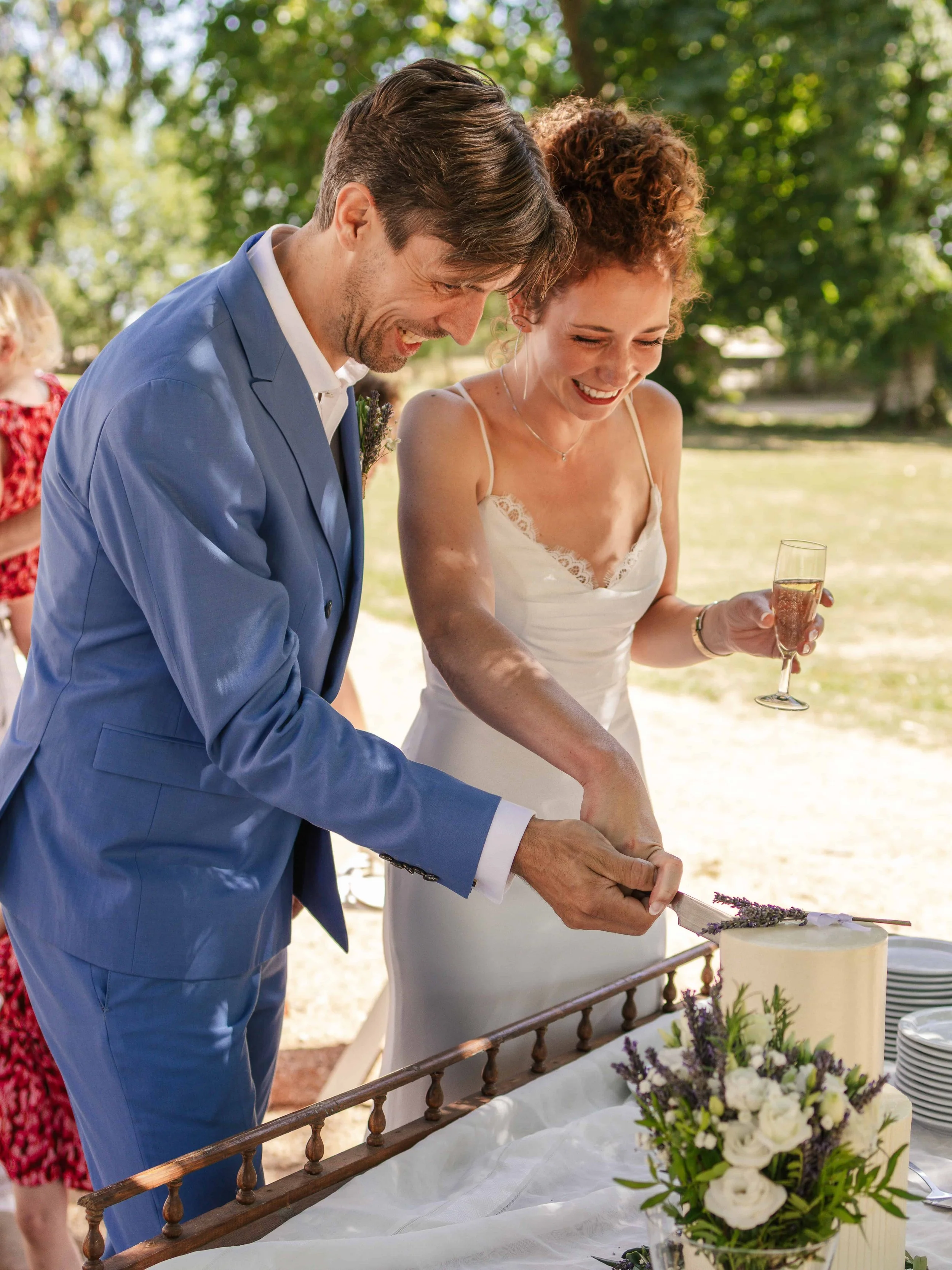 Een koppel, een man in een blauwe pak en een vrouw in een witte jurk, snijdt samen een bruidstaart op een buitenbruiloft. Ze glimlachen terwijl ze de taart snijden en een glas champagne vasthouden.