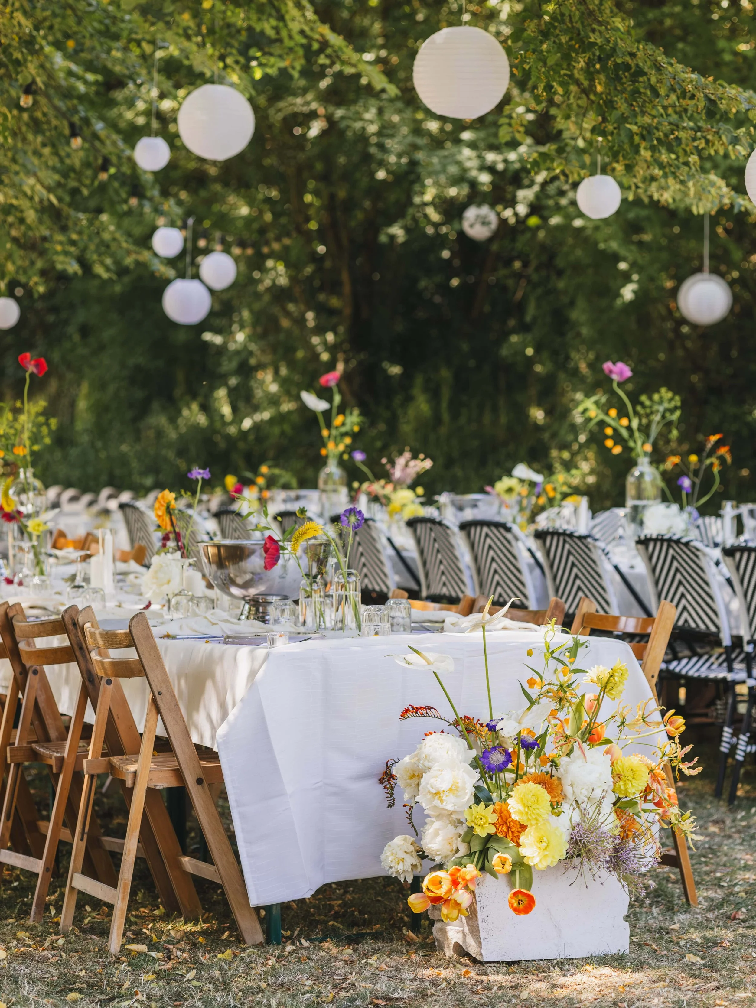 Een prachtig gedekte buiten eettafel met witte tafelkleed, kleurrijke bloemen en glazen, omgeven door bomen en lantaarns.
