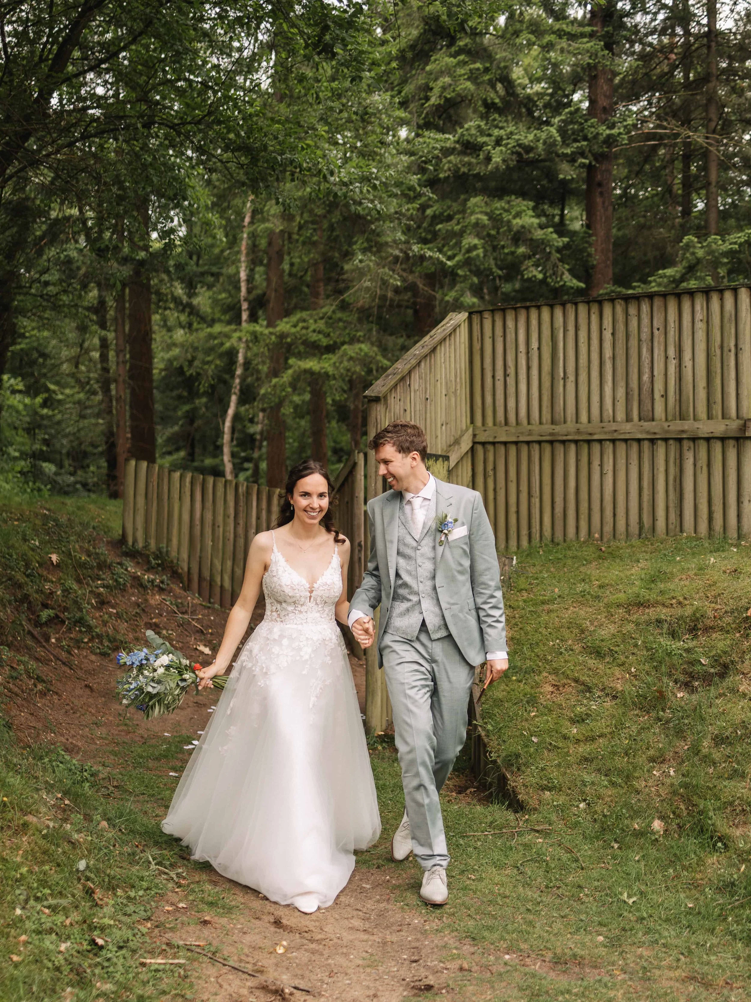 Een pasgetrouwd stel wandelt hand in hand door een bosrijke omgeving op hun trouwdag.