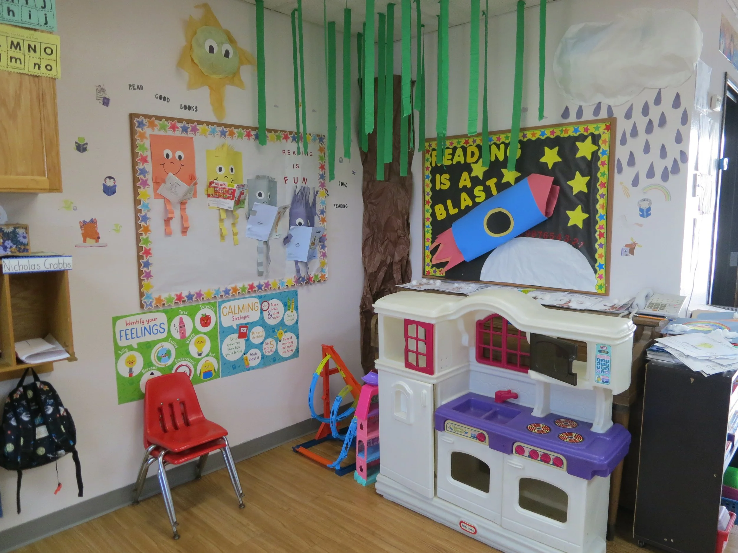 Decorated preschool classroom corner with colorful posters, a red chair, a toy kitchen set, and wall decorations including a smiling sun and paper rain clouds. (The Cross Schools of Education - Walnut, CA)