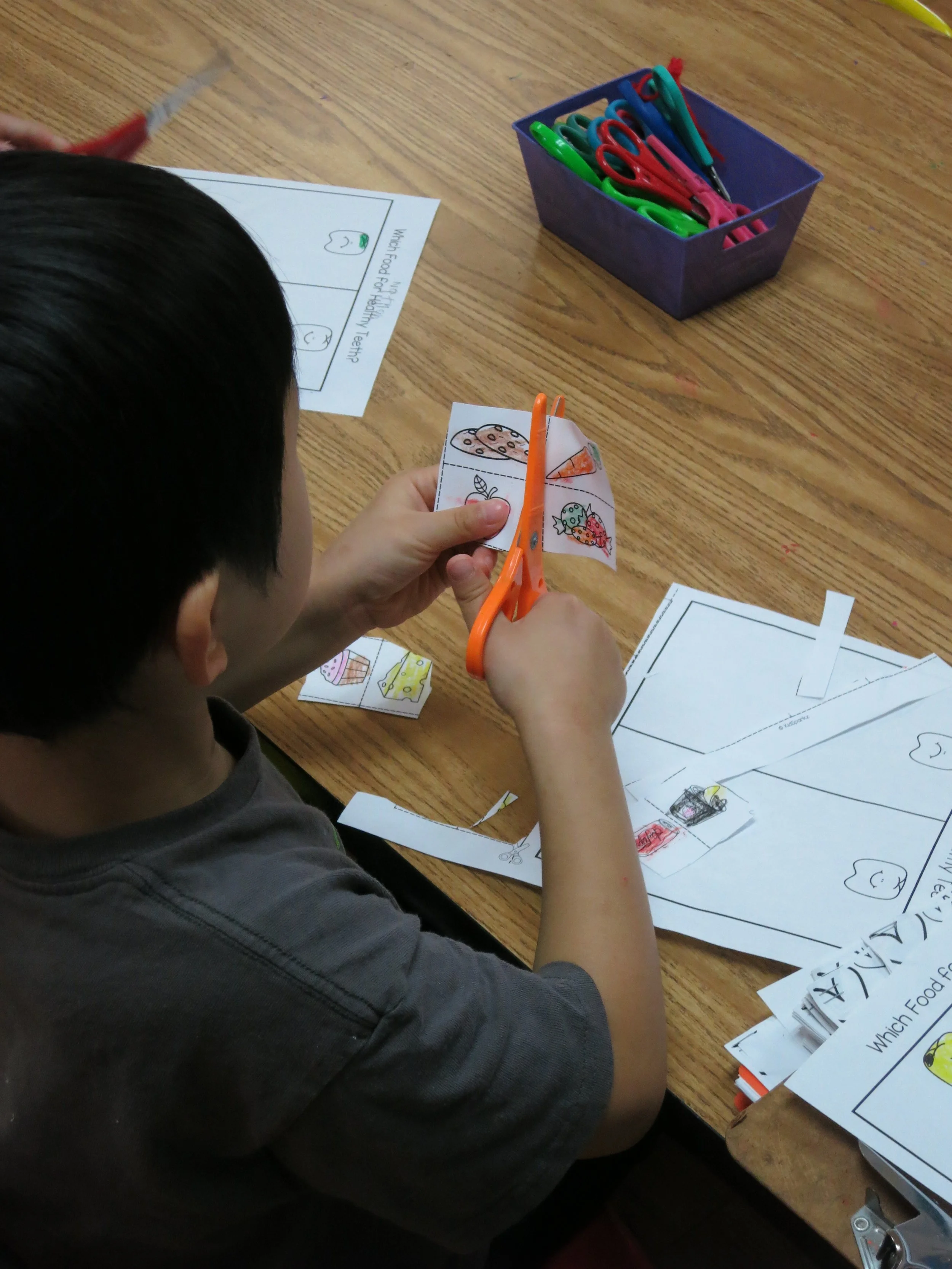 A preschool student using orange scissors to cut out a picture of a dessert from a coloring sheet, with coloring sheets, scissors, and a box of colorful scissors on a wooden table. (The Cross Schools of Education - Walnut, CA)