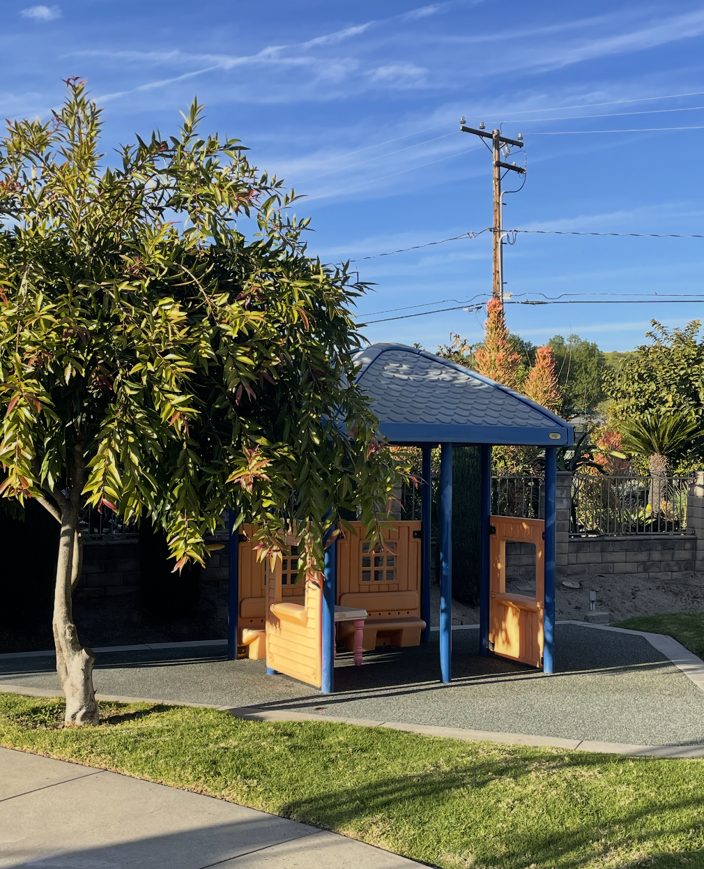 A small children's playhouse with a blue roof and orange walls, set in a park area with a large tree nearby, under a blue sky with some clouds.  (The Cross Schools of Education - Walnut, CA)