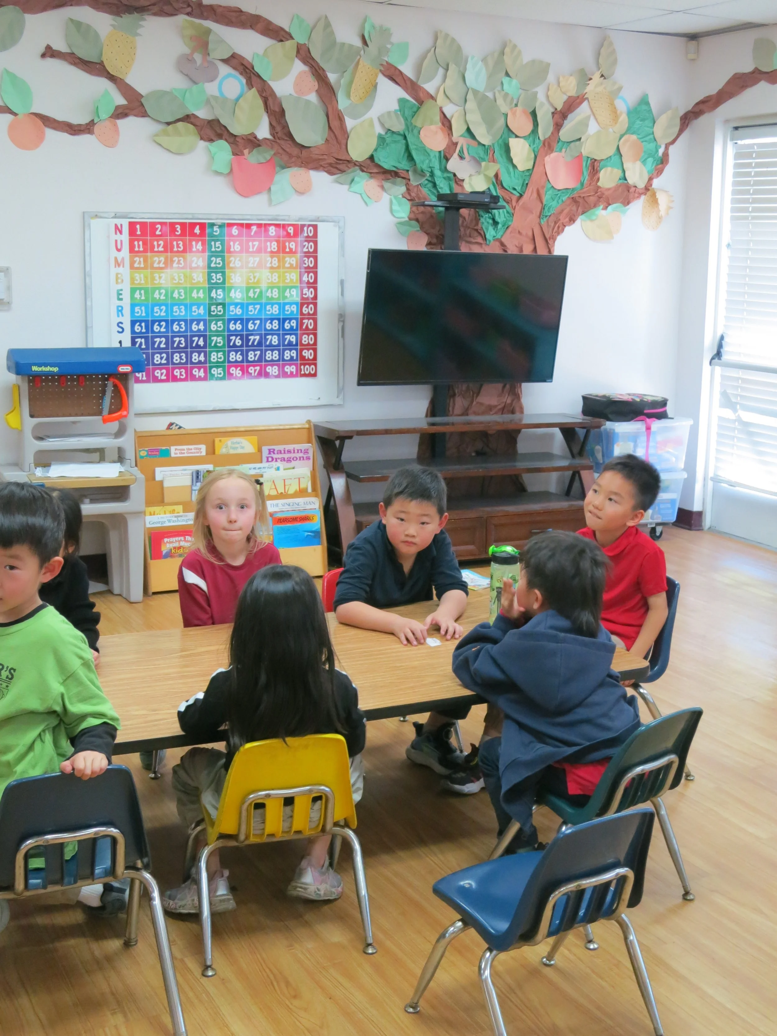 A private kindergarten classroom with children sitting around a table, with a large paper tree with leaves and fruit on the wall behind them, and a colorful number chart. There is also a television mounted on a stand and some shelves with books and s
