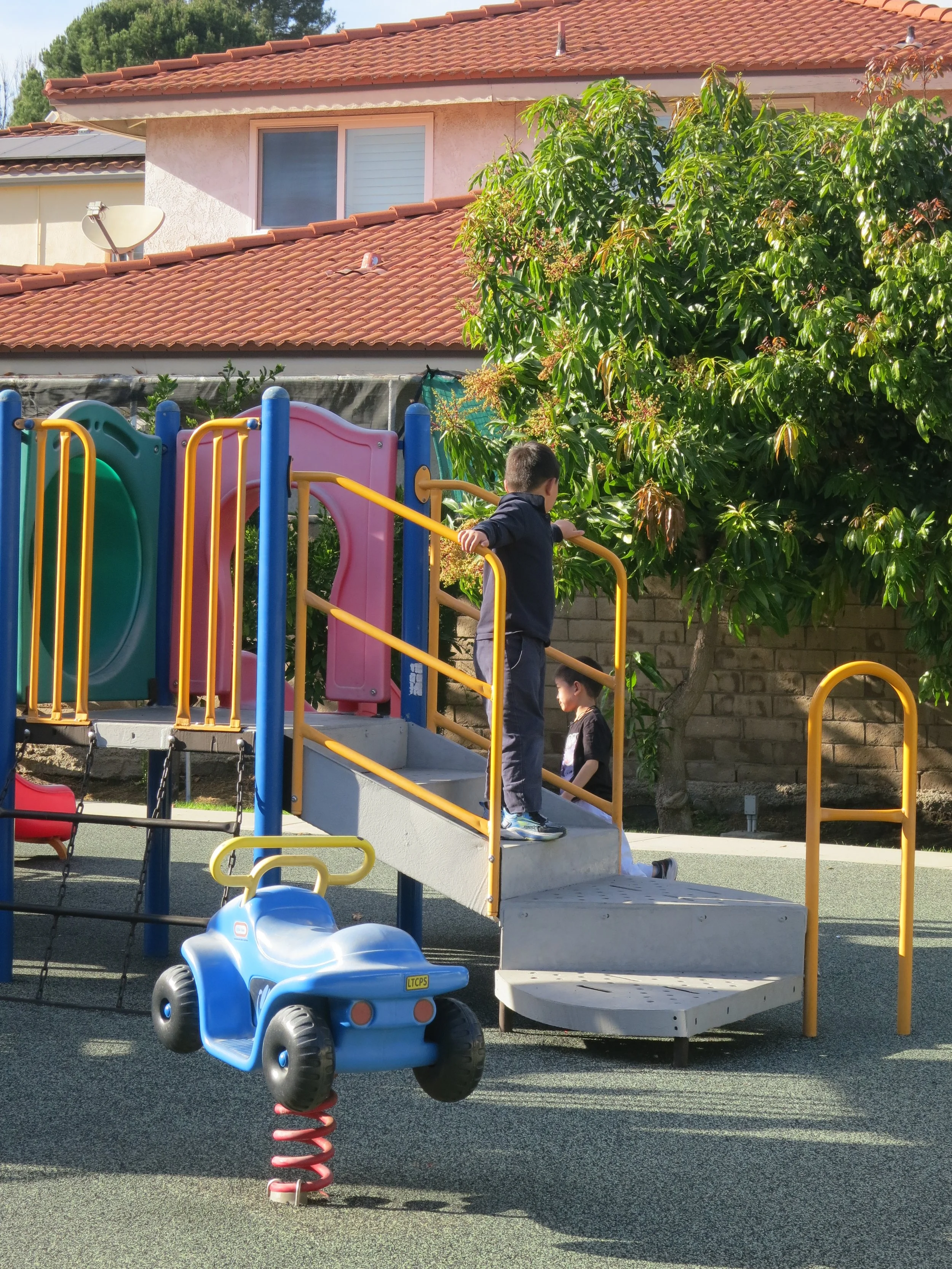 Children playing on a colorful playground with slides and a spring rider shaped like a car, near a house with a red-tiled roof and green trees. (The Cross Schools of Education - Walnut, CA)