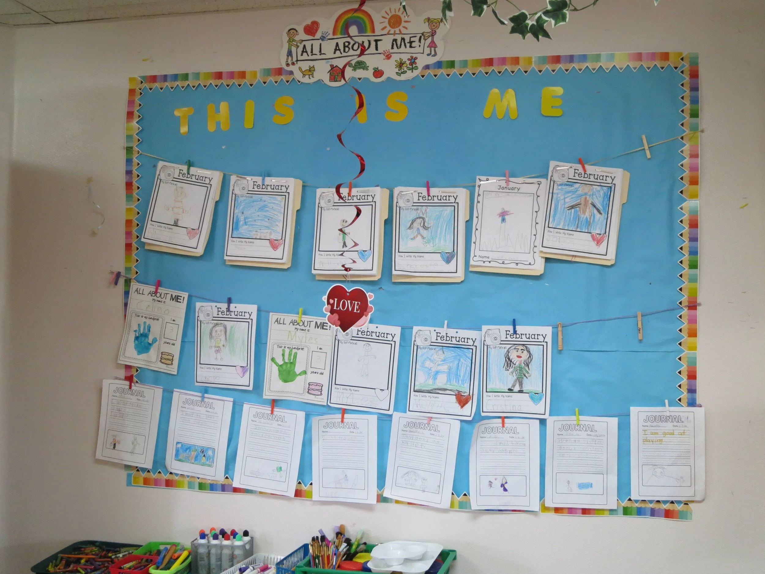 Preschool classroom bulletin board titled "All About Me" with children's drawings, writings, and journal entries about themselves for February and January, decorated with colorful borders and a red heart with the word "Love." (The Cross Schools of Ed