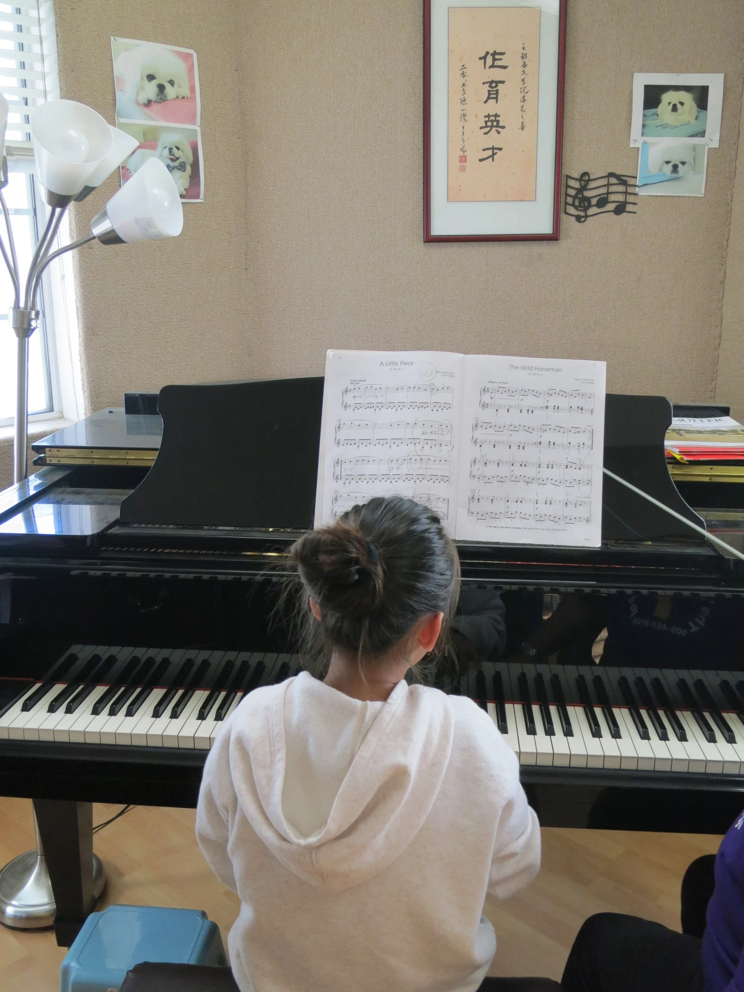 A girl with her hair in a bun seated at a black piano, practicing sheet music titled "A Little Piece" and "The Wild Horseman." The room has beige walls decorated with pictures of a white dog, a framed piece of Asian calligraphy, and a musical note wa