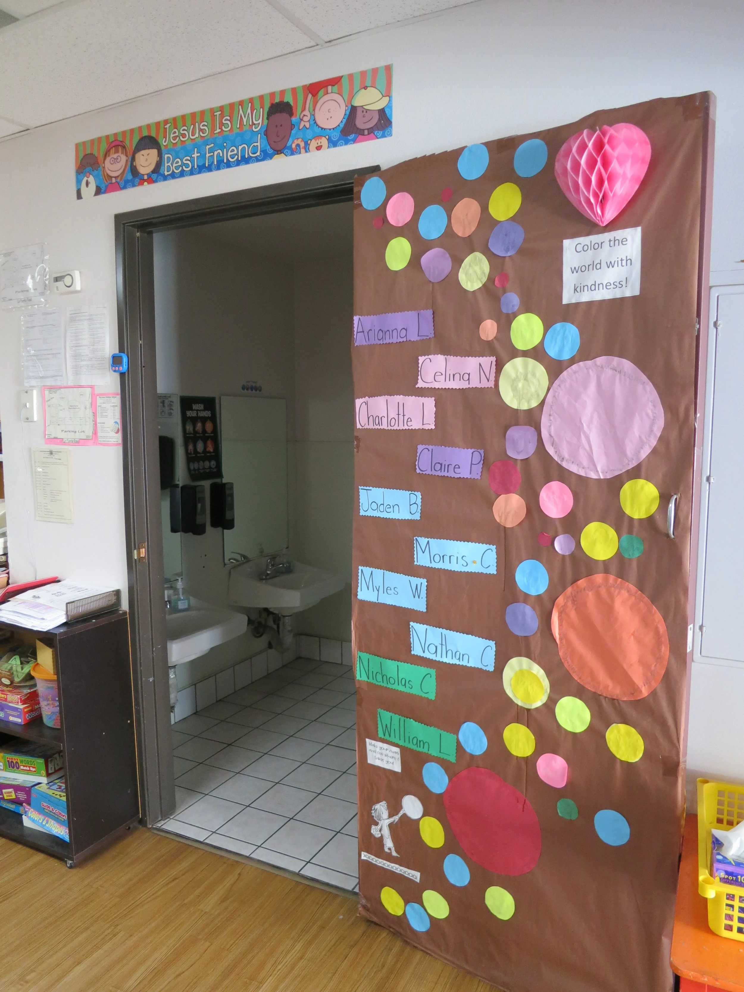 Colorful classroom bulletin board with names and circles, some labeled with students’ names, and a pink honeycomb heart. A sign says, 'Color the world with kindness!' (The Cross Schools of Education - Walnut, CA)