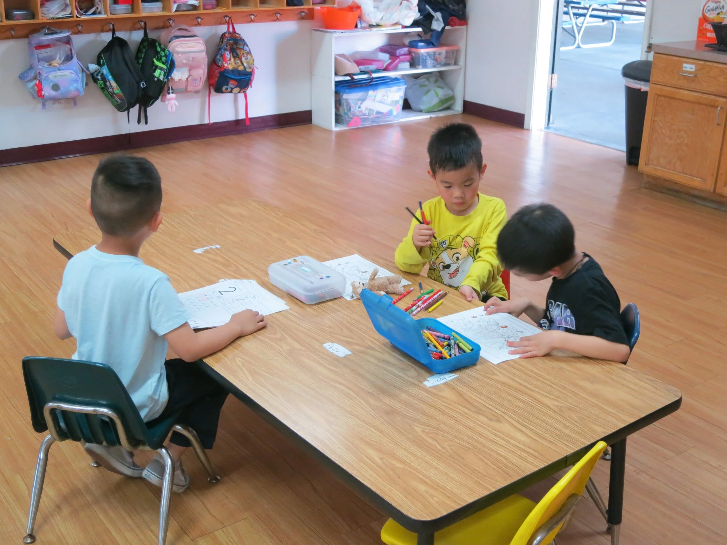 Three preschoolers sit at a wooden table in a classroom, coloring and drawing with markers and crayons. Backpack hooks with colorful backpacks are on the wall, and storage shelves with supplies are in the background. An open door reveals an outdoor a