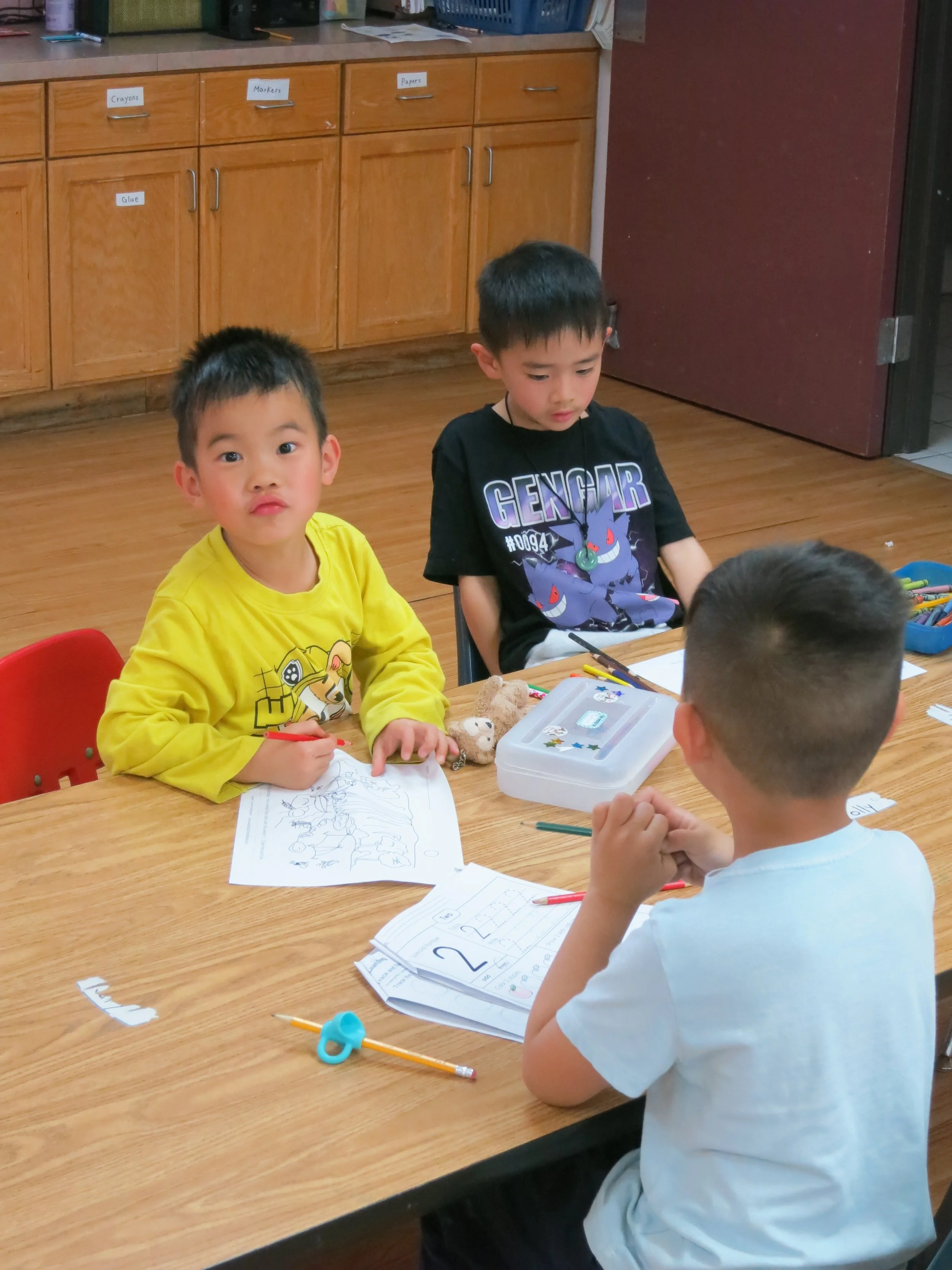 Three private Christian kindergarten boys are sitting at a wooden table in a classroom, working on coloring and numbers with papers, markers, and toys. (The Cross Schools of Education - Walnut, CA)
