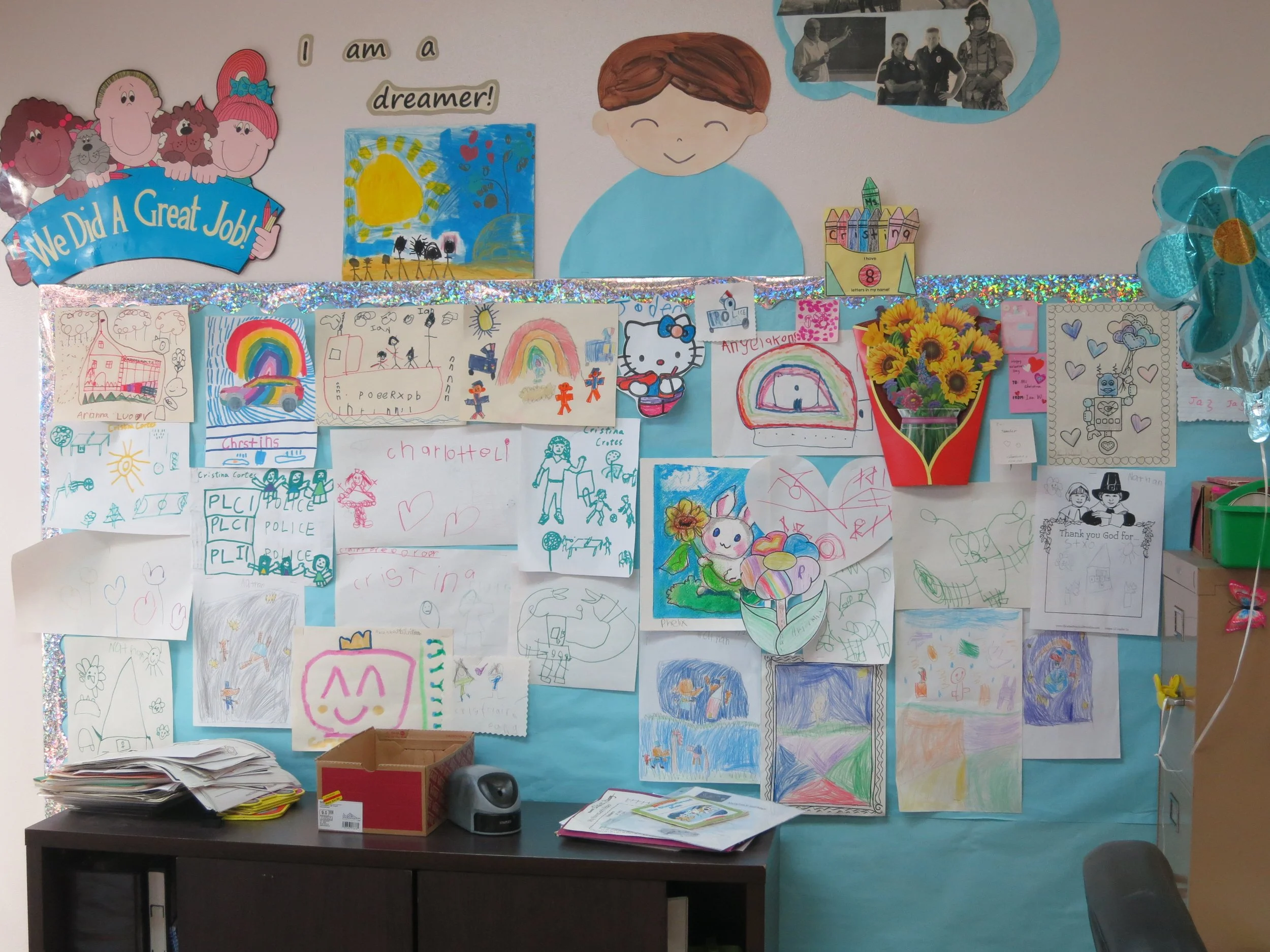 Classroom bulletin board decorated with children's drawings, colorful artworks, and posters. Some drawings include rainbows, houses, and animals. There is a banner at the top with cartoon characters and the words "We Did A Great Job!" A box, some pap