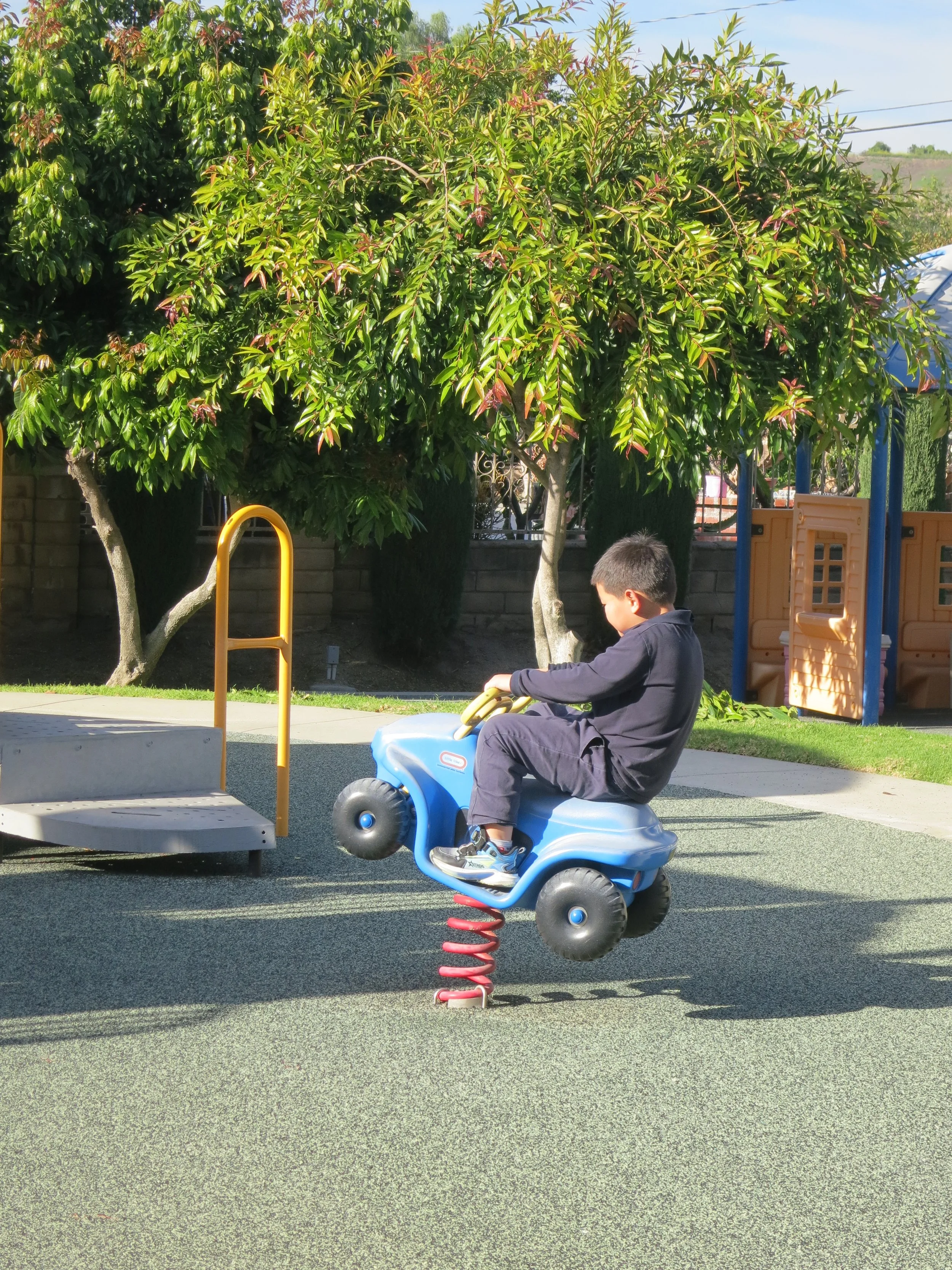 A young boy is sitting on a blue spring rocker at a playground, with a tree and playground equipment in the background. (The Cross Schools of Education - Walnut, CA)