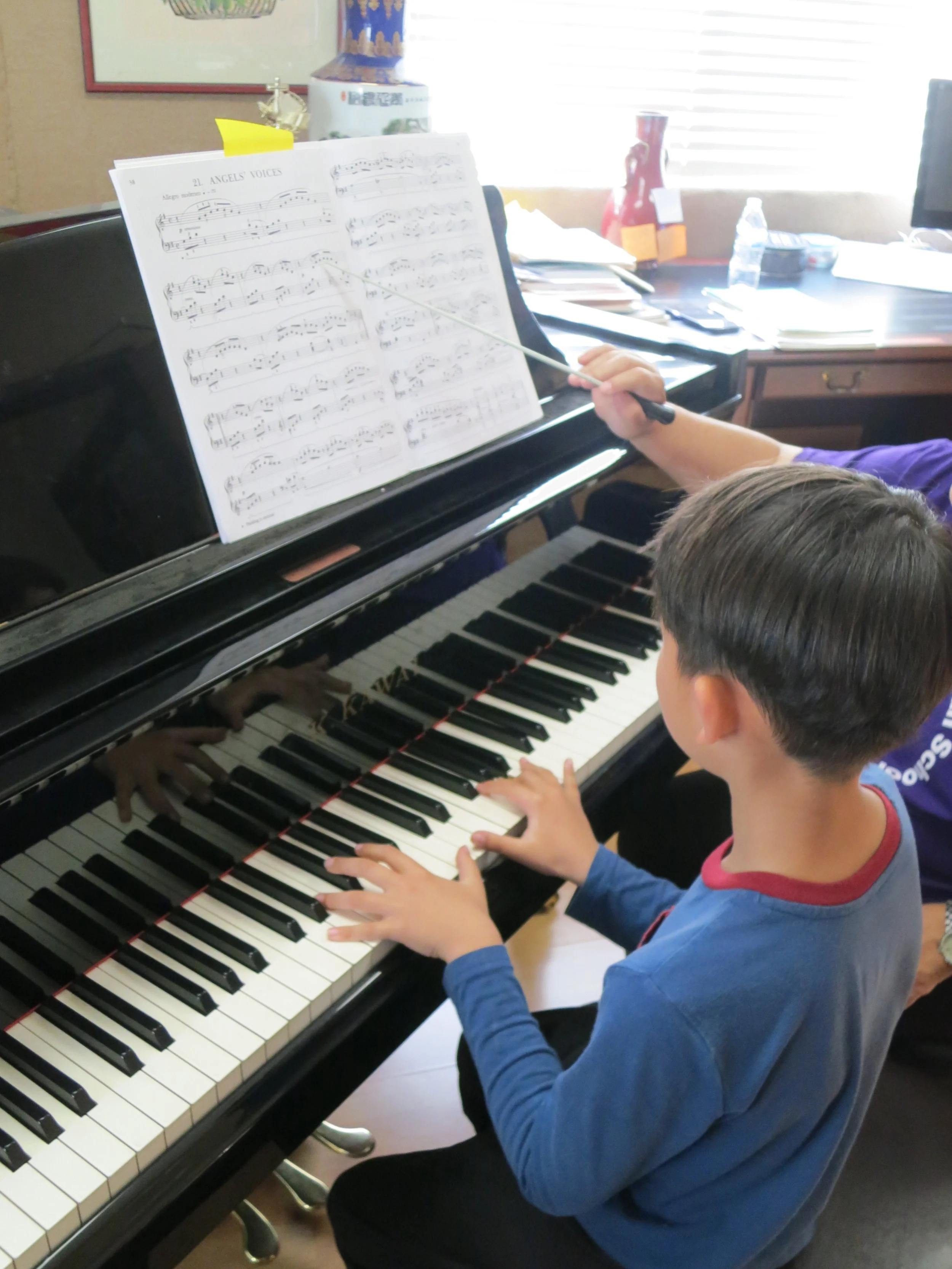 A young boy playing the piano with a teacher guiding him, as indicated by her hand holding the piano baton. Sheet music titled 'Angel's Voices' is on the music stand in front of them. The room has a desk with papers, water bottles, and some decorativ