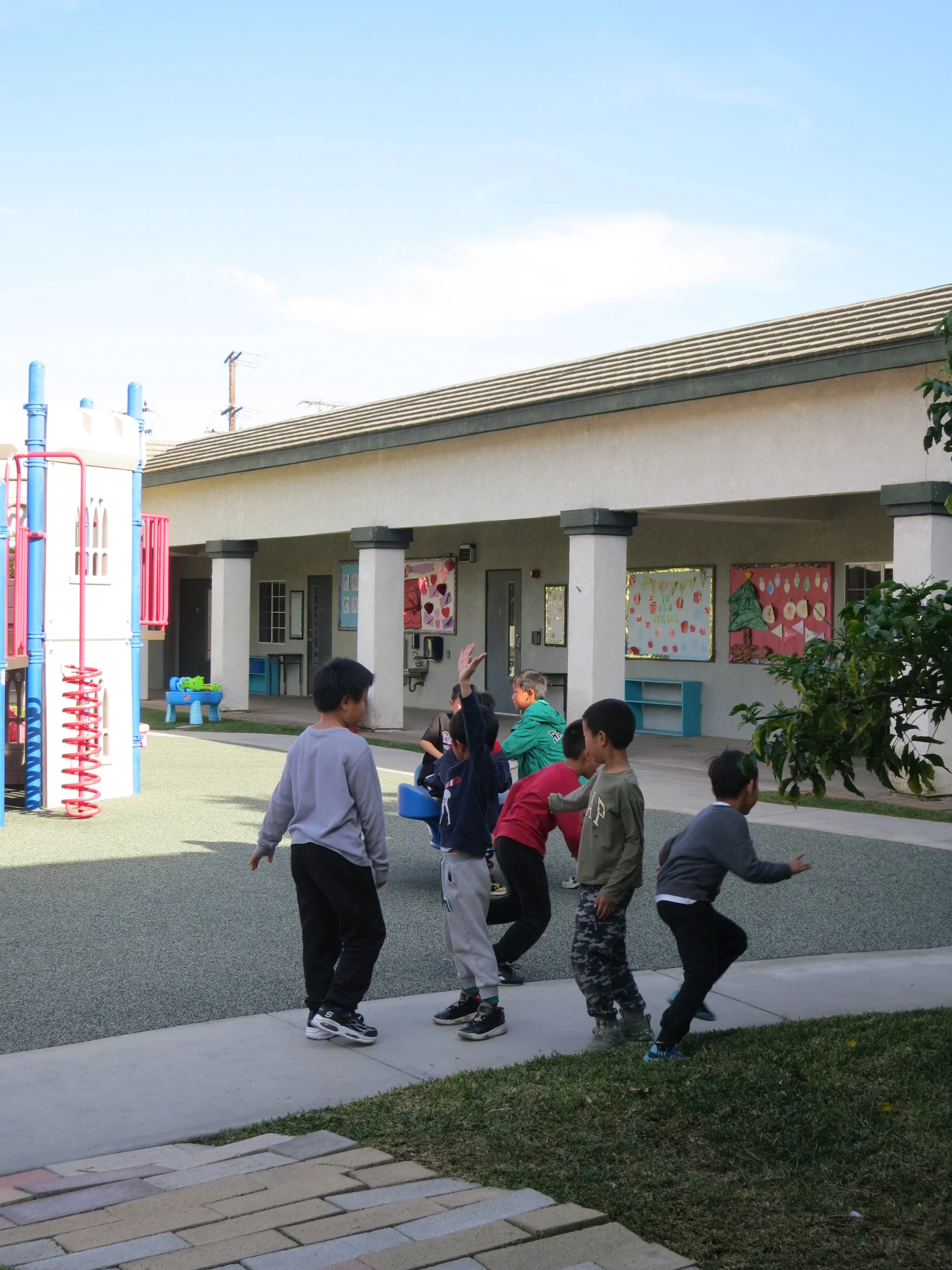 A group of after-school program children playing outdoors on a playground in front of a school building, some kids standing and others crouching or jumping, with colorful artwork on the walls. (The Cross Schools of Education - Walnut, CA)