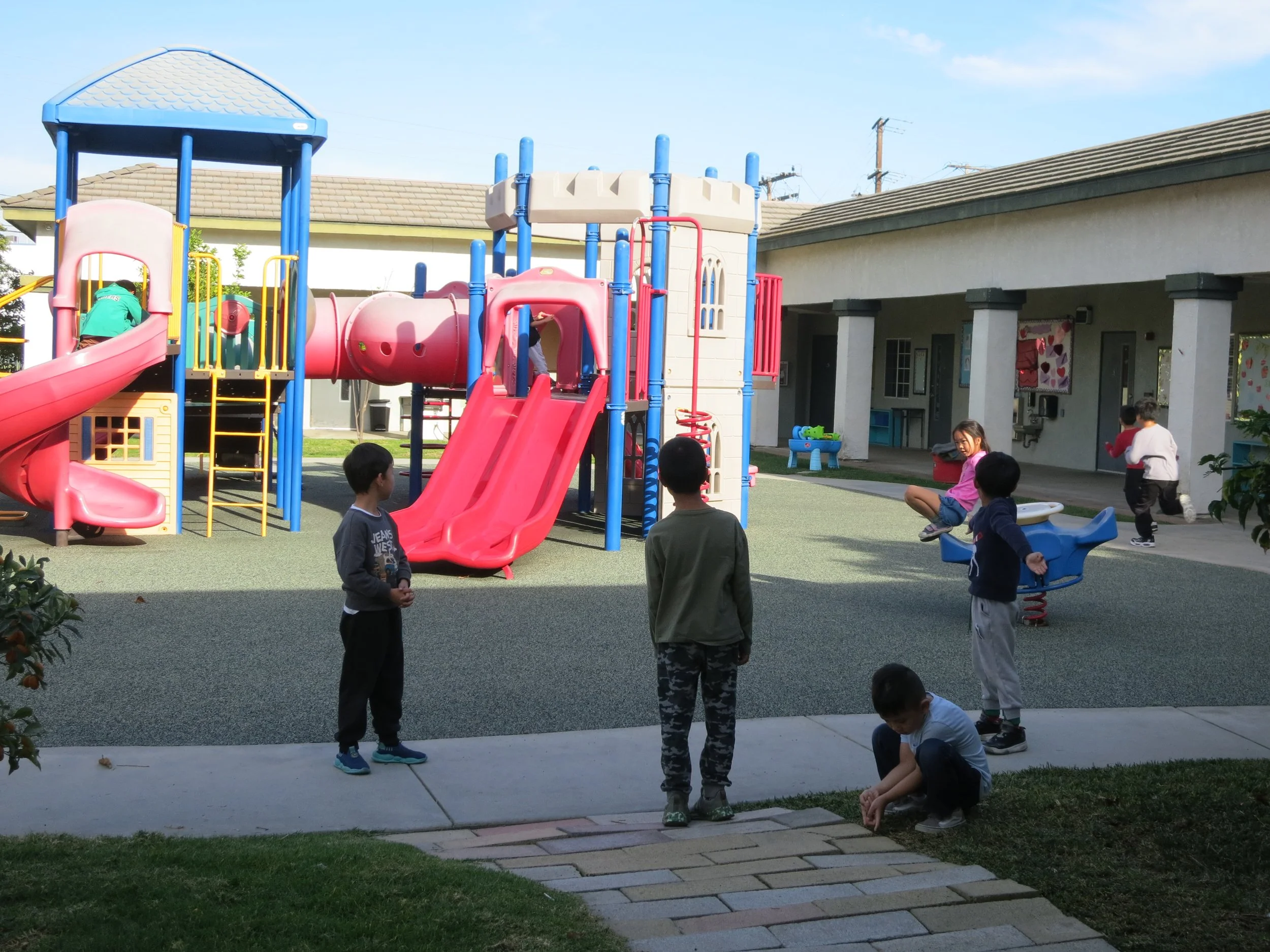 Children playing on playground equipment including slides and see-saw in an outdoor playground area, with a building in the background. (The Cross Schools of Education - Walnut, CA)