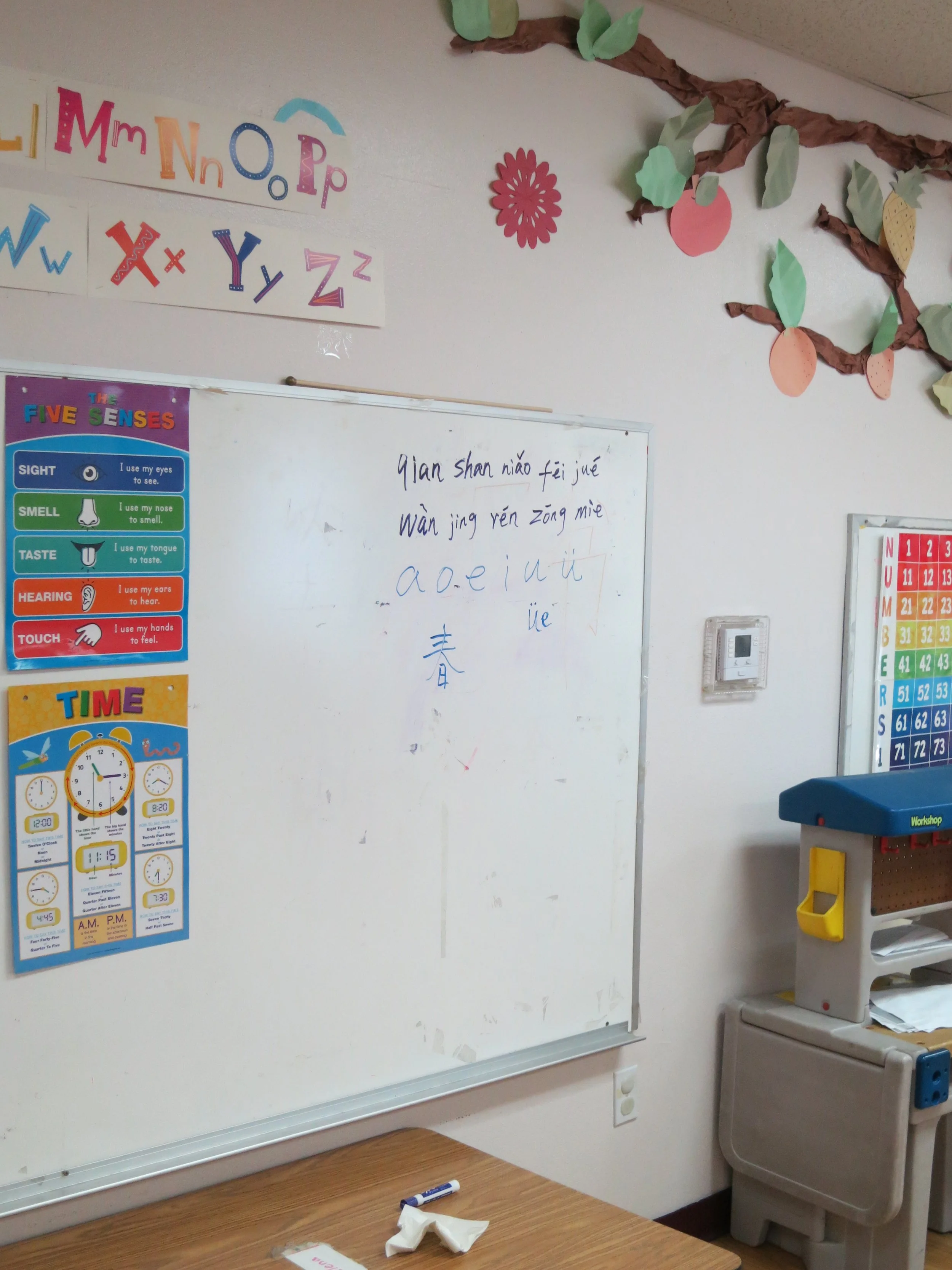 Inside a classroom, a whiteboard with writing in Chinese and Pinyin, educational posters about the five senses and time, and colorful decorations of plants and fruits on the wall. (The Cross Schools of Education - Walnut, CA)